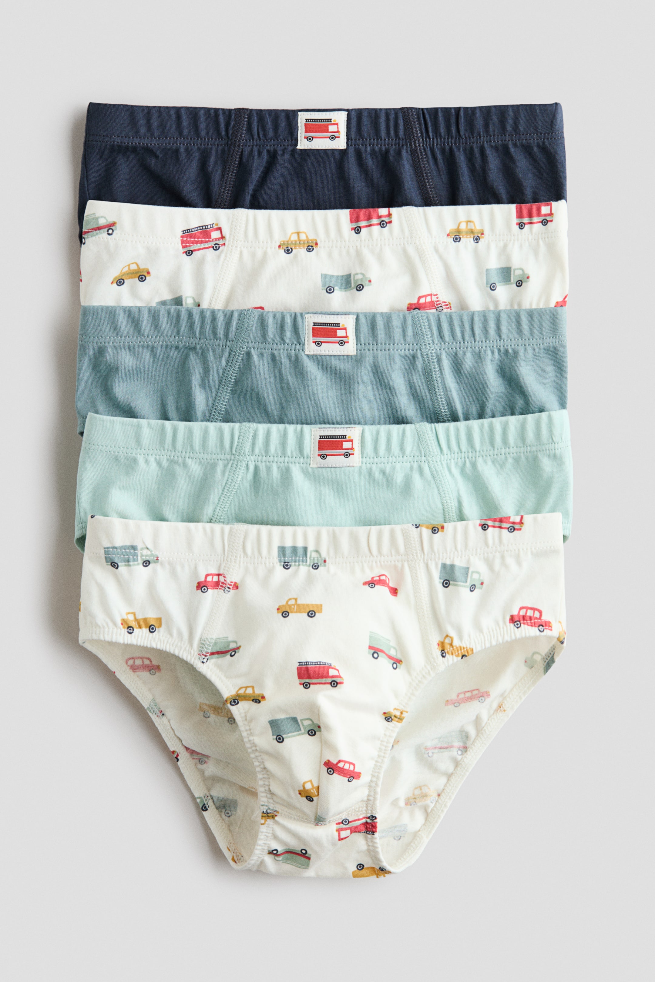 View larger image: A 5-pack of cotton boys' briefs is showcased, with five briefs stacked and slightly overlapping, each displaying its front view. The stack features a navy blue brief at the top, followed by an off-white brief with colorful vehicle patterns, then a light blue brief, a light mint green brief, and finally another off-white brief with colorful vehicle patterns at the bottom. Each brief includes an elastic waistband and a small rectangular tag with a red fire truck graphic on the front.