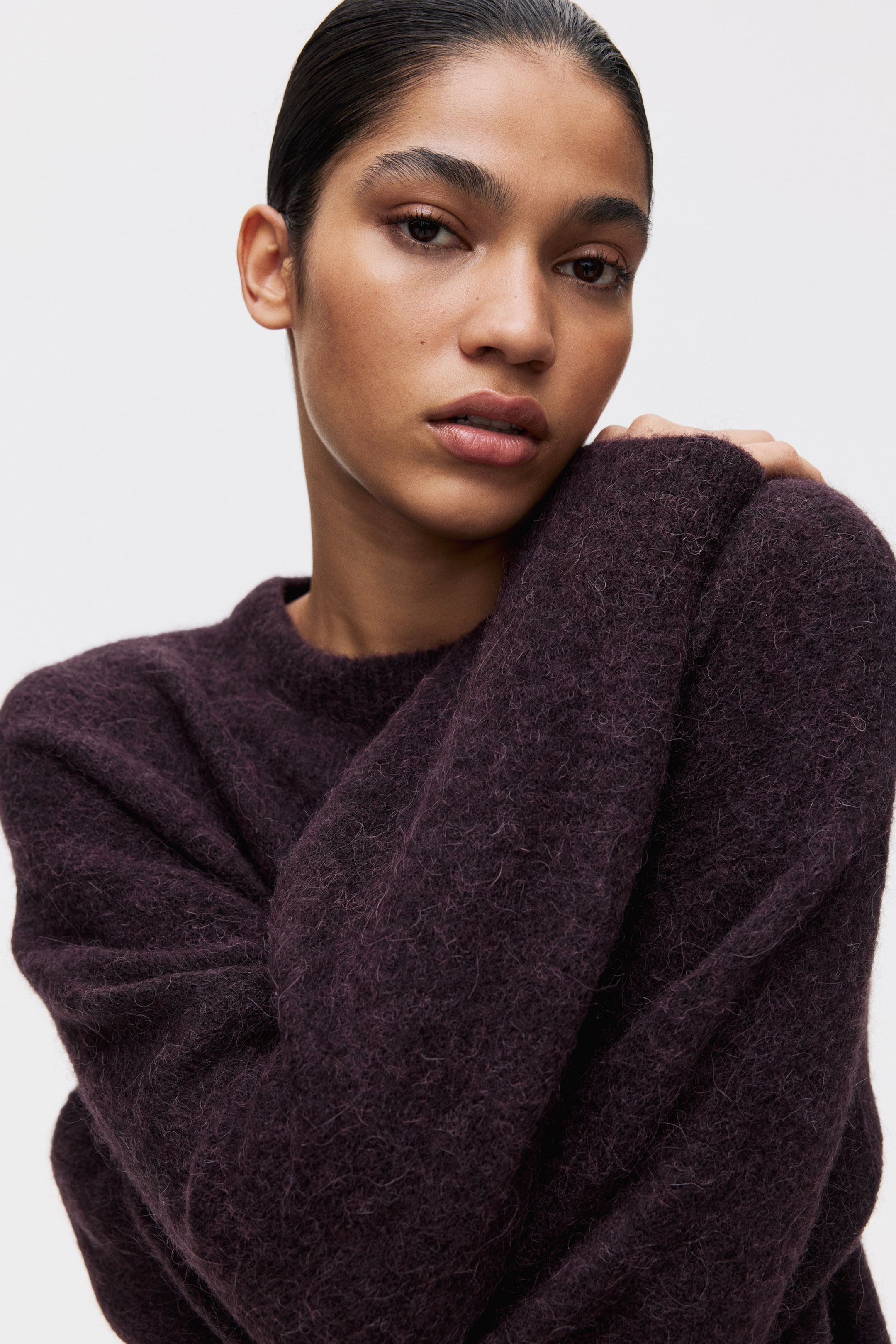 Ampliar la imagen: Se lleva puesto un suéter de cuello redondo color púrpura oscuro, con una textura suave y peluda de tejido visible. Una manga se sostiene sobre el pecho, mostrando el ajuste relajado y la caída del suéter.