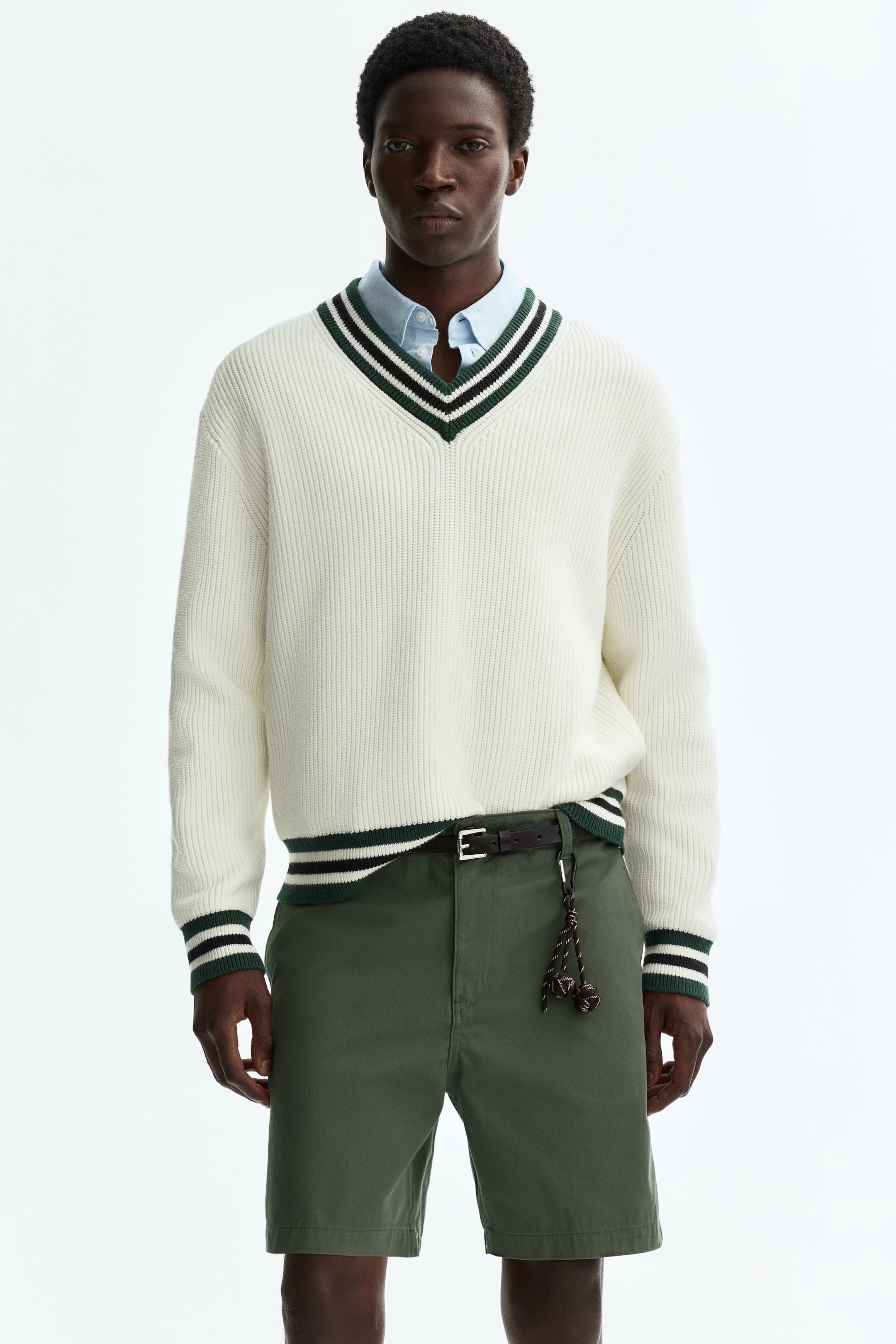 View larger image: The off-white ribbed knit V-neck jumper showcases green, black, and white stripes along its V-neck, hem, and cuffs. A light blue collared shirt is visible beneath the jumper.