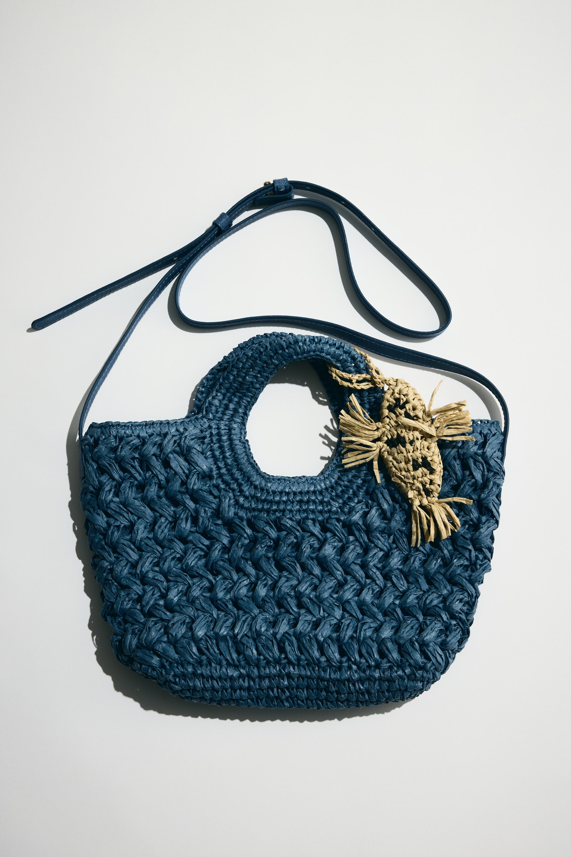 Ampliar la imagen: Un bolso cruzado de paja de papel trenzado azul muestra su parte frontal, con dos asas redondeadas en la parte superior. Un dije en forma de pez de color marrón claro cuelga del frente, y una delgada correa ajustable para el hombro en azul es visible extendiéndose desde los lados.