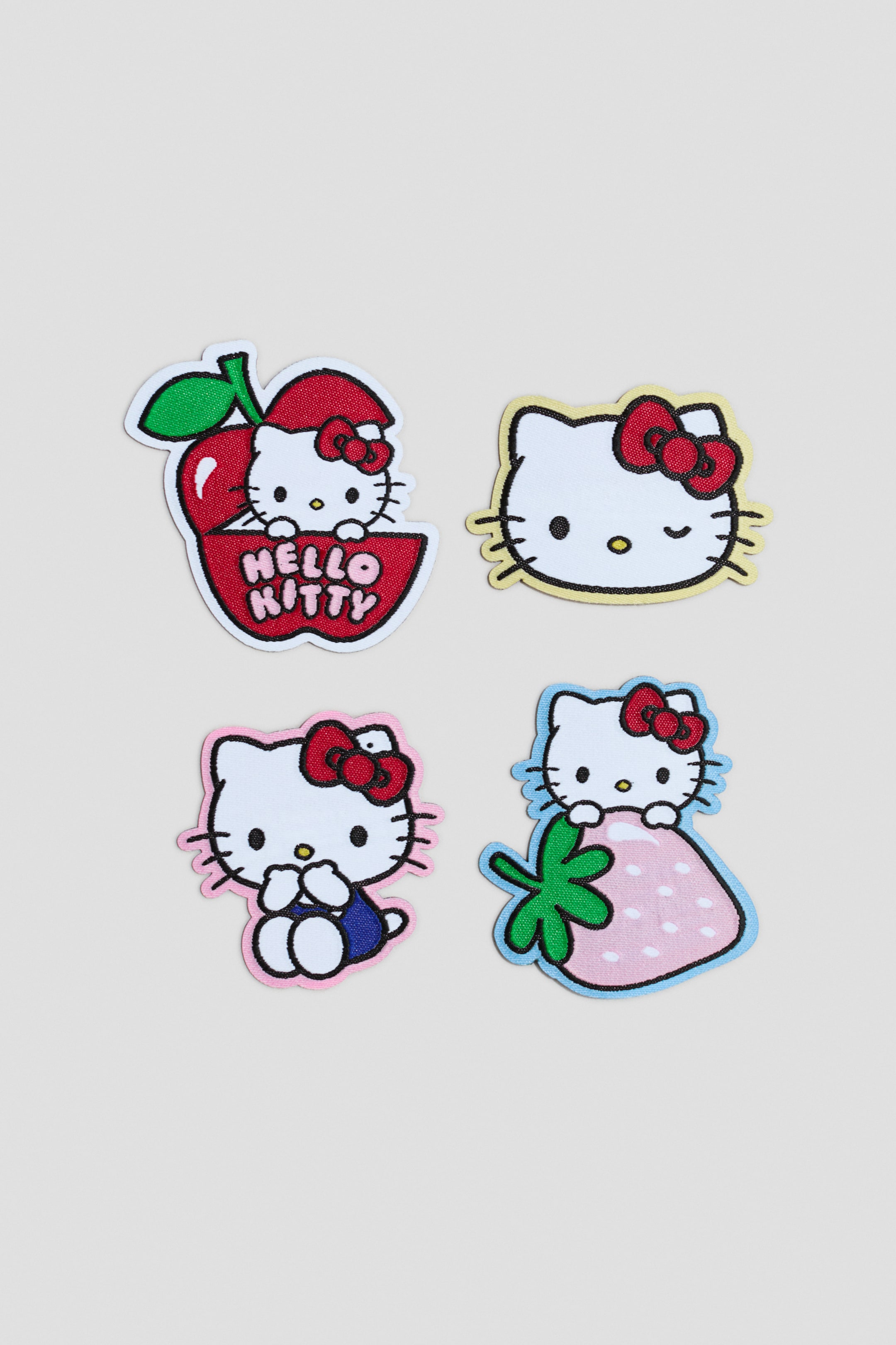 Ver imagem maior: Os remendos de reparação apresentam quatro designs distintos bordados da Hello Kitty. Cada remendo exibe a Hello Kitty numa pose diferente ou com um acessório distinto: espreitando de uma maçã vermelha com o texto "HELLO KITTY", piscando o olho, sentada com um traje azul, e espreitando por cima de um morango cor-de-rosa.