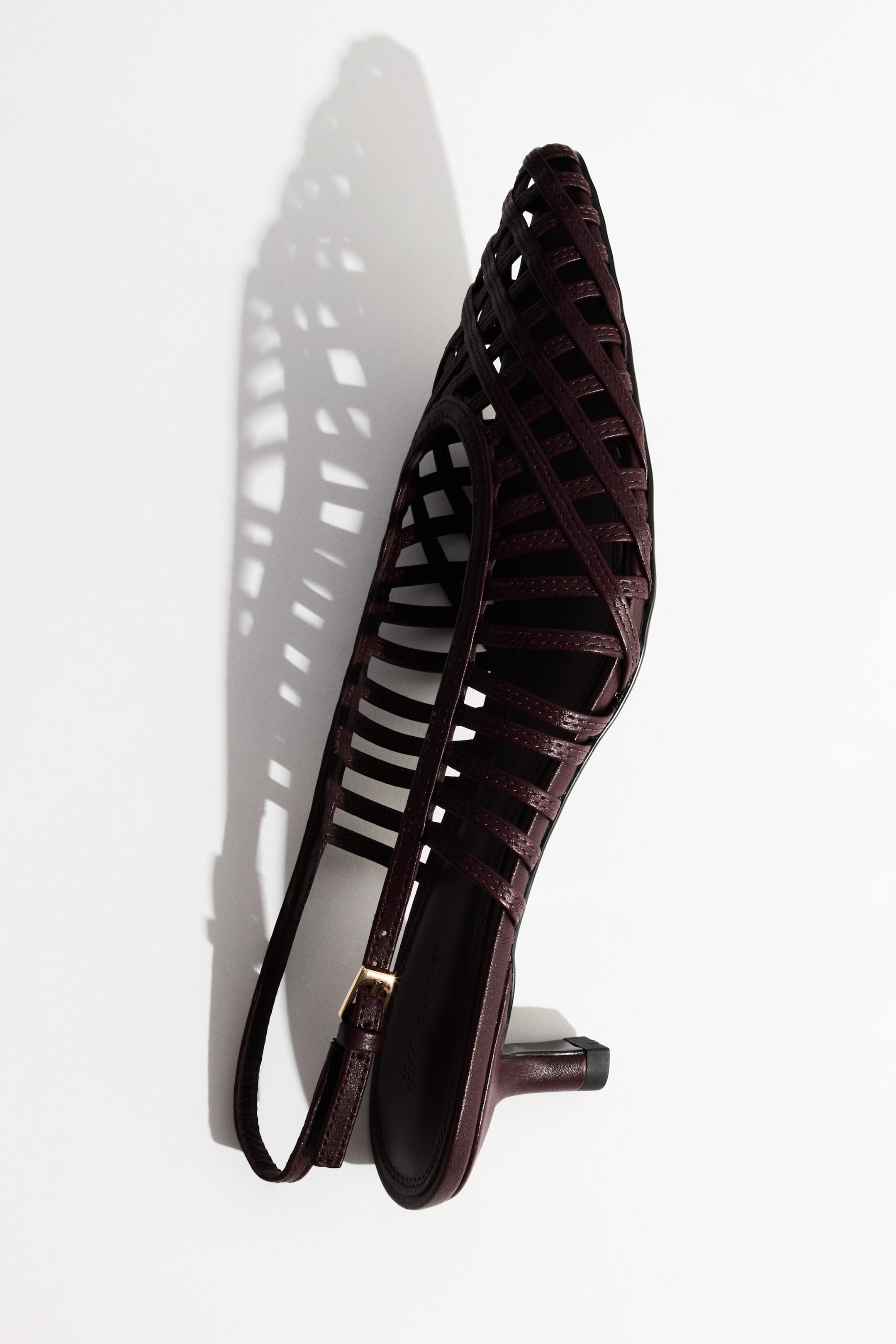 View larger image: A solitary dark burgundy slingback pump rests on its side, positioned diagonally, with its pointed toe directed upwards and to the left. It showcases a woven lattice upper design, a low flared kitten heel, and a slender slingback strap adorned with a gold-toned buckle.