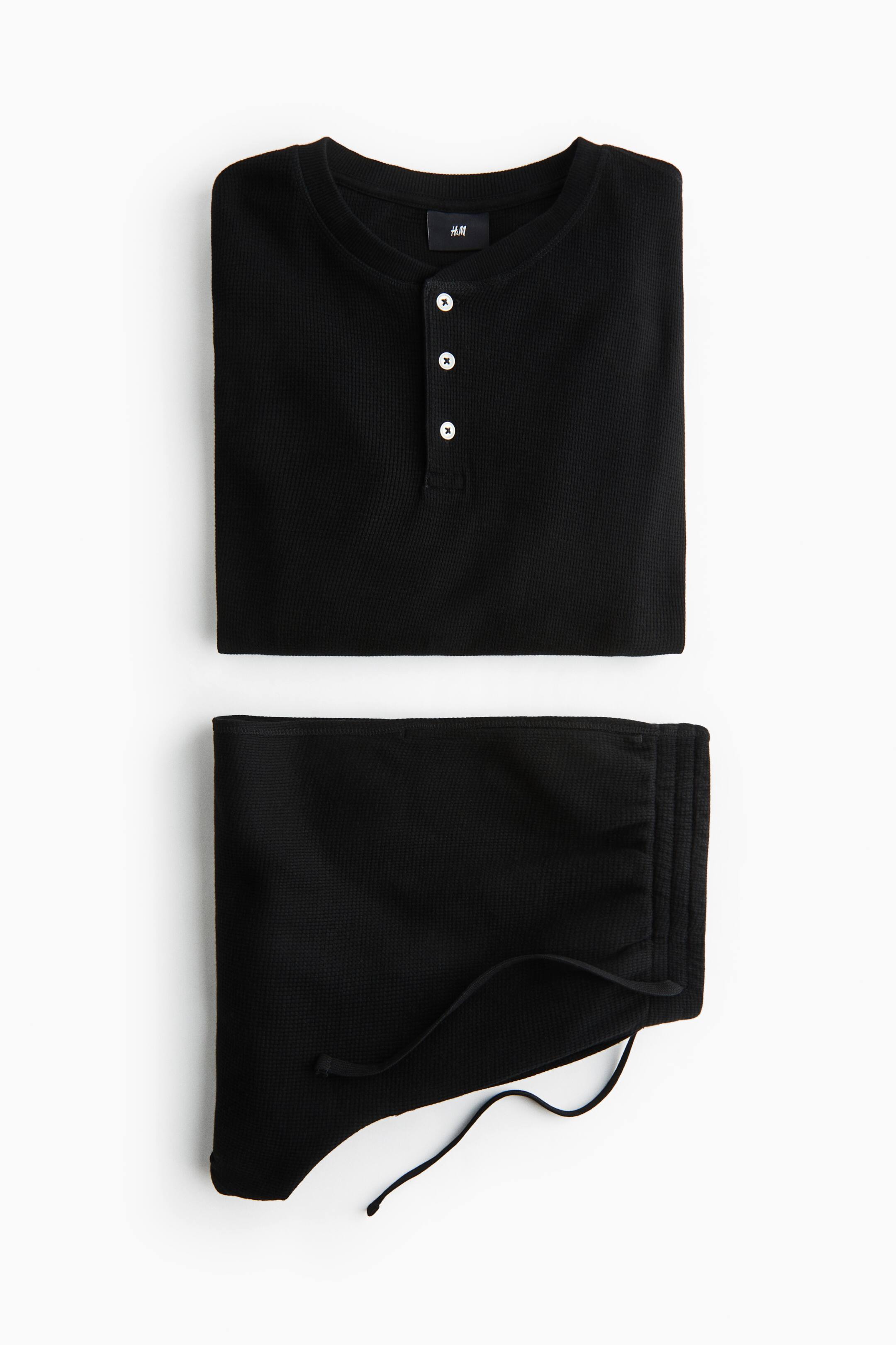 Agrandir l'image: Un ensemble thermique noir en maille gaufrée comprend une chemise Henley à col rond avec patte de boutonnage à trois boutons et boutons blancs, ainsi qu’un pantalon assorti présentant un détail côtelé et un cordon de serrage noir ; les deux pièces sont pliées à plat.