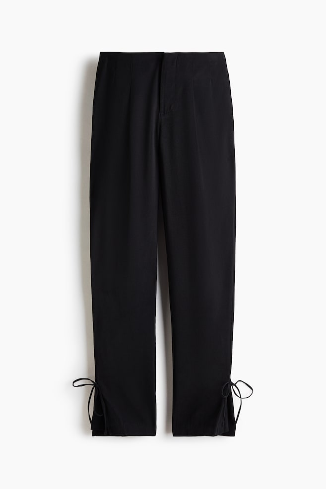 These black trousers are shown from the front, featuring a straight-leg silhouette with subtle front pleats and a clean, minimalist waistband. The design is detailed with adjustable self-tie drawstrings forming delicate bows at the outer ankle cuffs. - 1
