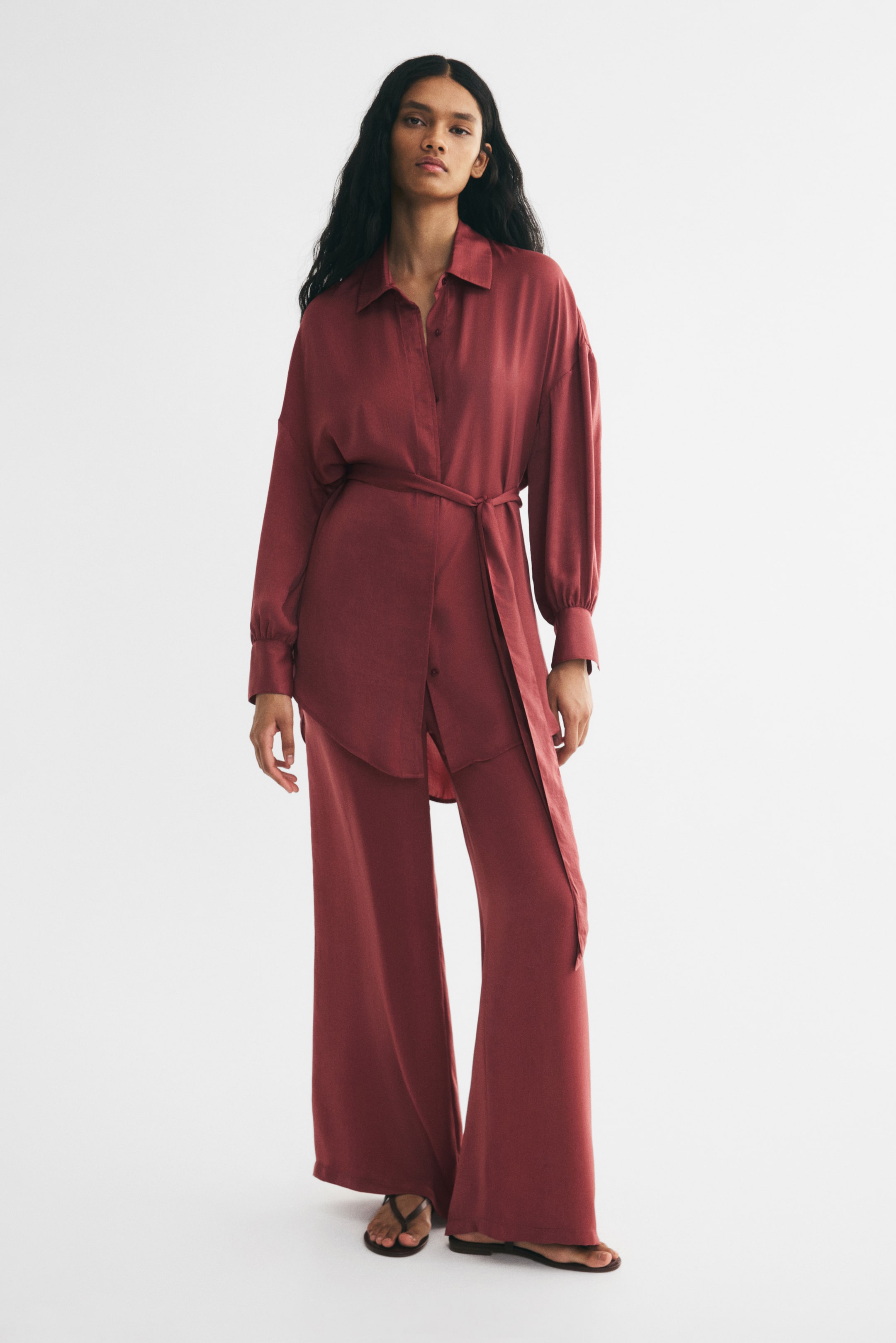 View larger image: The model is wearing trousers in a maroon gently crinkled weave with a slight sheen. They feature a wide-leg silhouette and full-length fit, styled with a matching long-sleeved shirt layered over the waist.