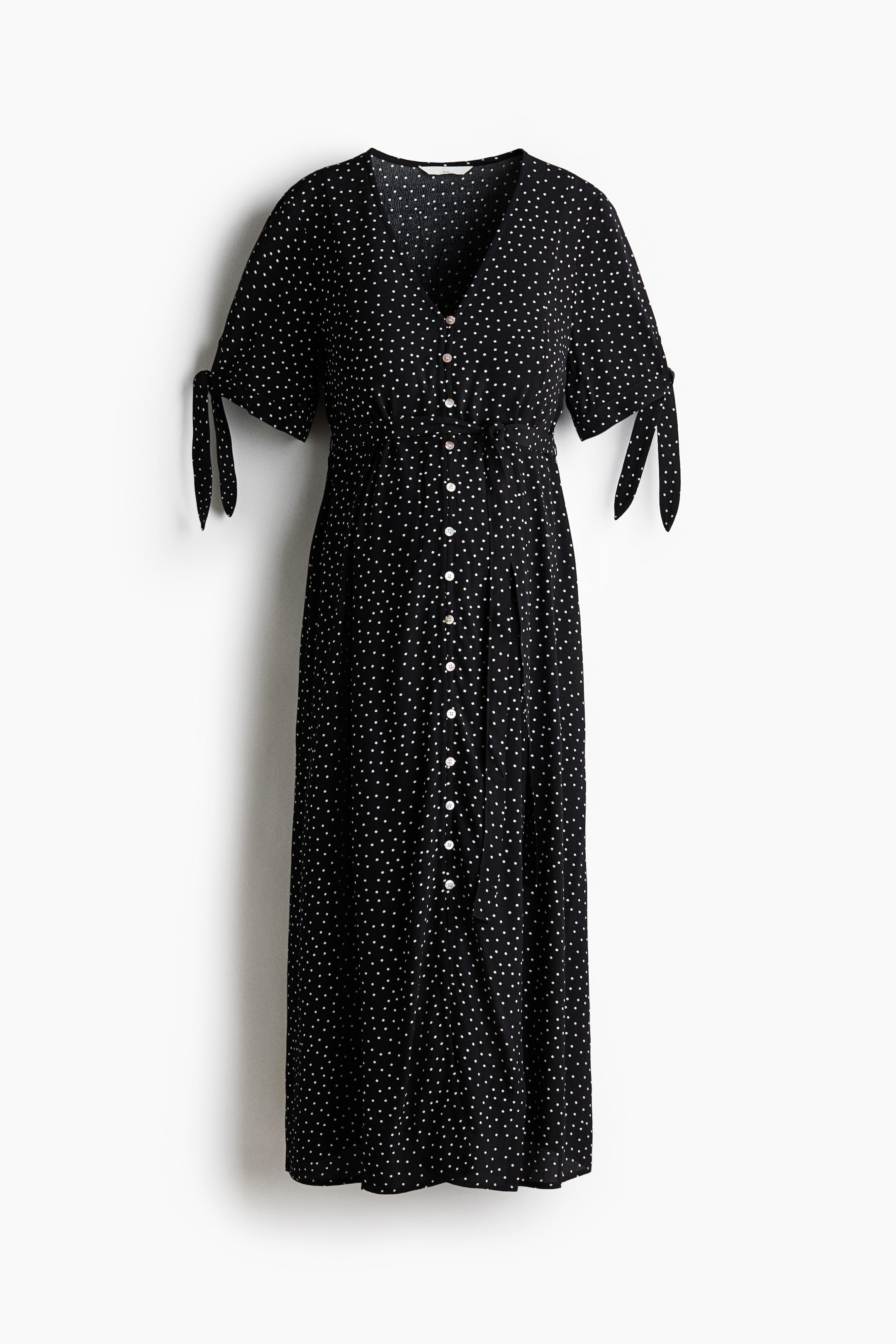View larger image: A black midi dress with white polka dots is shown from the front, featuring a V-neckline and short sleeves with tie details. It displays a full button-down front with light-colored buttons and a cinched waist.