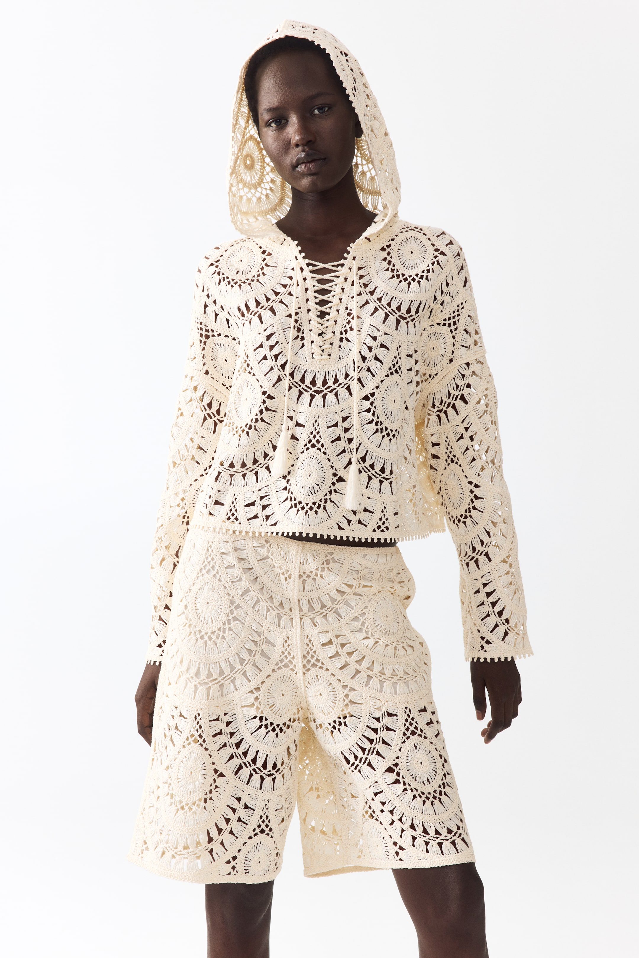 View larger image: A cream-colored crochet matching set showcases a long-sleeved hooded top paired with knee-length shorts, both facing forward. The top features a lace-up V-neckline with tasseled drawstrings and an intricate openwork circular pattern that extends onto the hood and shorts, with all hems finished in a bobble trim.