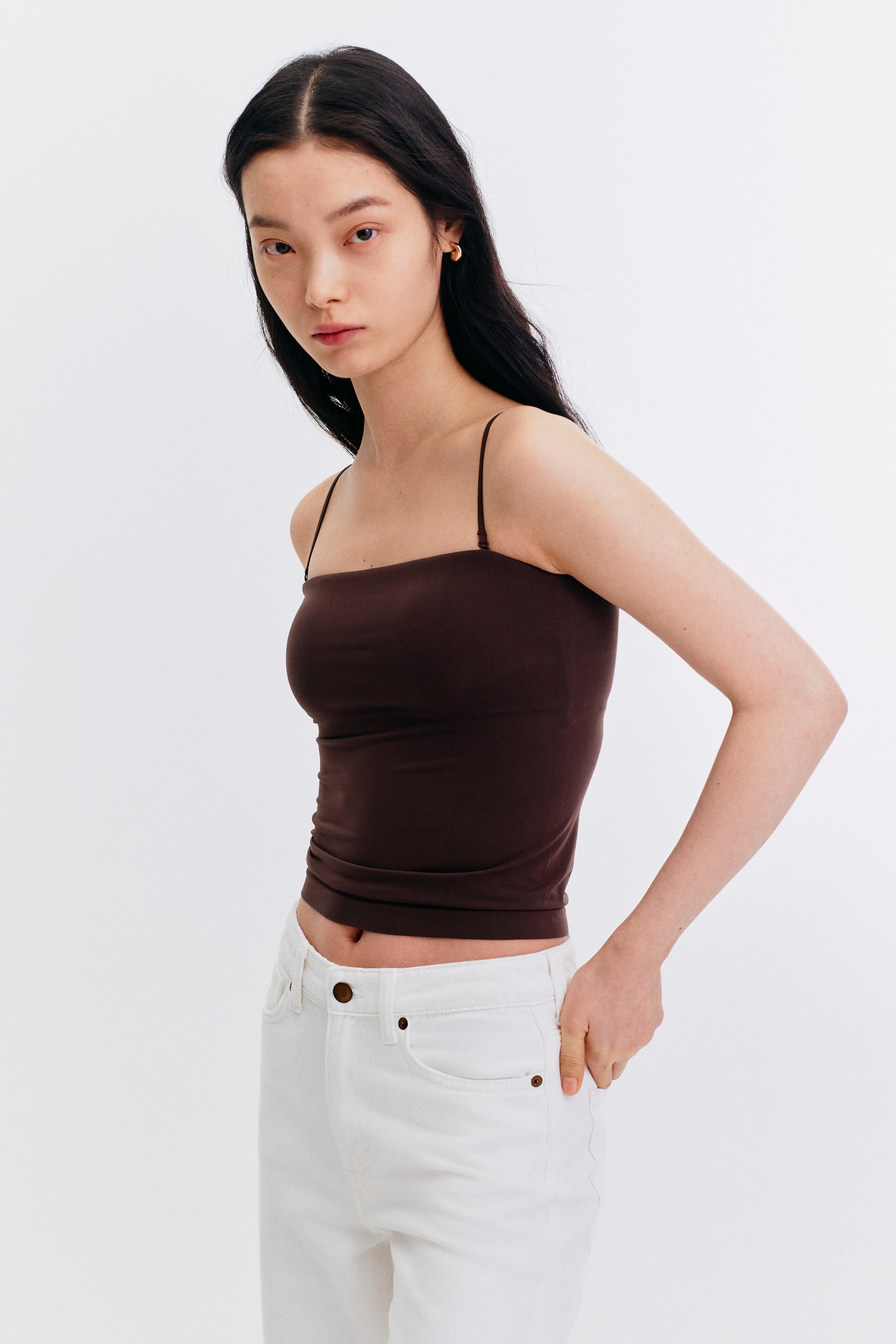 View larger image: The model is wearing a dark brown camisole top featuring a square neckline and slender spaghetti straps. It's paired with white jeans, with the model casually placing her hand in the front pocket of the jeans.