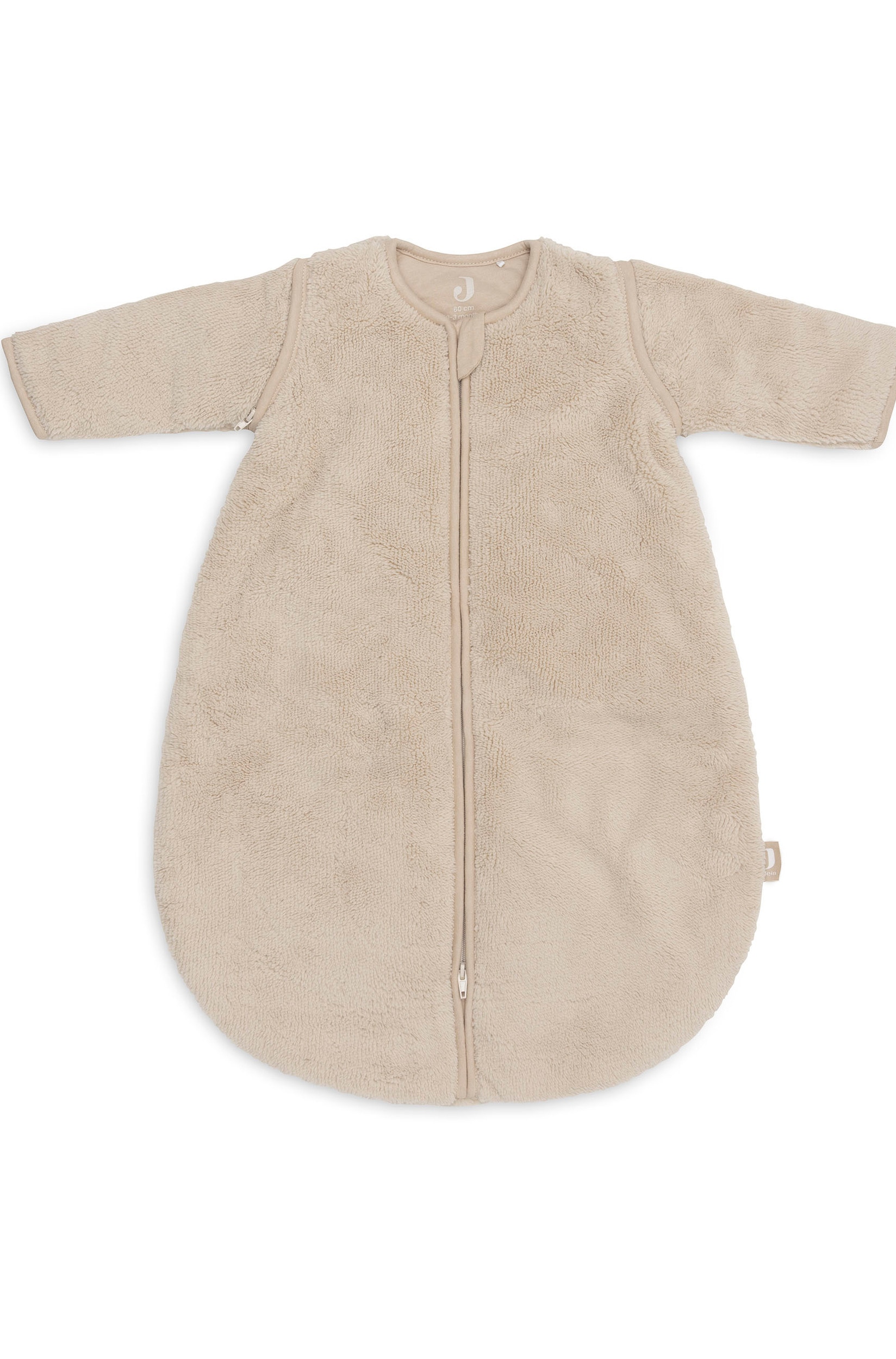 Een beige, zachte babyslaapzak met lange mouwen hangt naar voren gericht aan een houten standaard en toont de zachte fleece-structuur en een ritssluiting in het midden over de volledige lengte. Aan de bovenkant van de ritssluiting hangt een klein, bijpassend zacht schaapje met een houten bijtring.