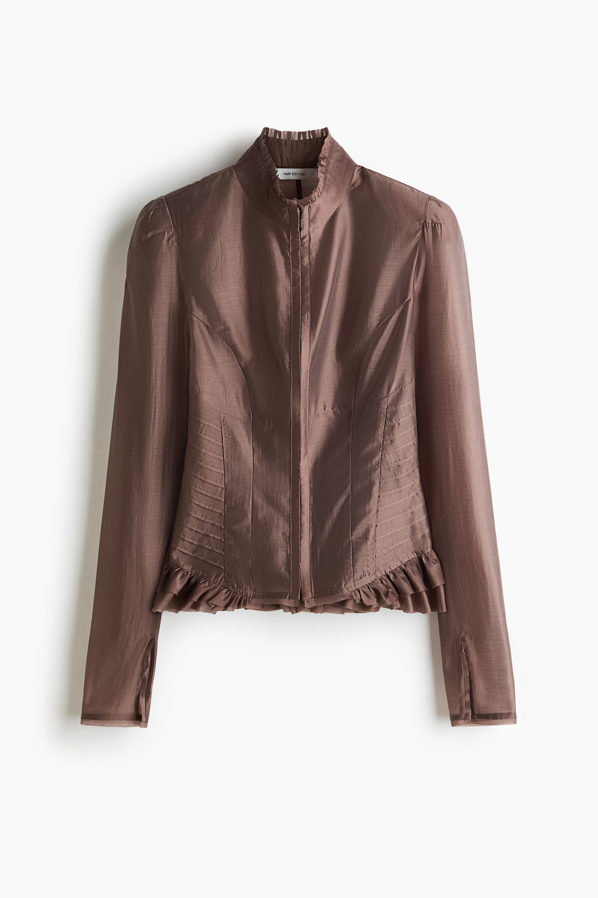 Une blouse marron ajustÃ©e, lÃ©gÃ¨rement transparente, avec un col montant, des Ã©paules structurÃ©es et des dÃ©tails plissÃ©s Ã  la taille est portÃ©e par le mannequin. La blouse est portÃ©e ouverte sur le devant, laissant apparaÃ®tre un collier pendentif dorÃ© et le ventre, et est associÃ©e Ã  un pantalon blanc taille haute.