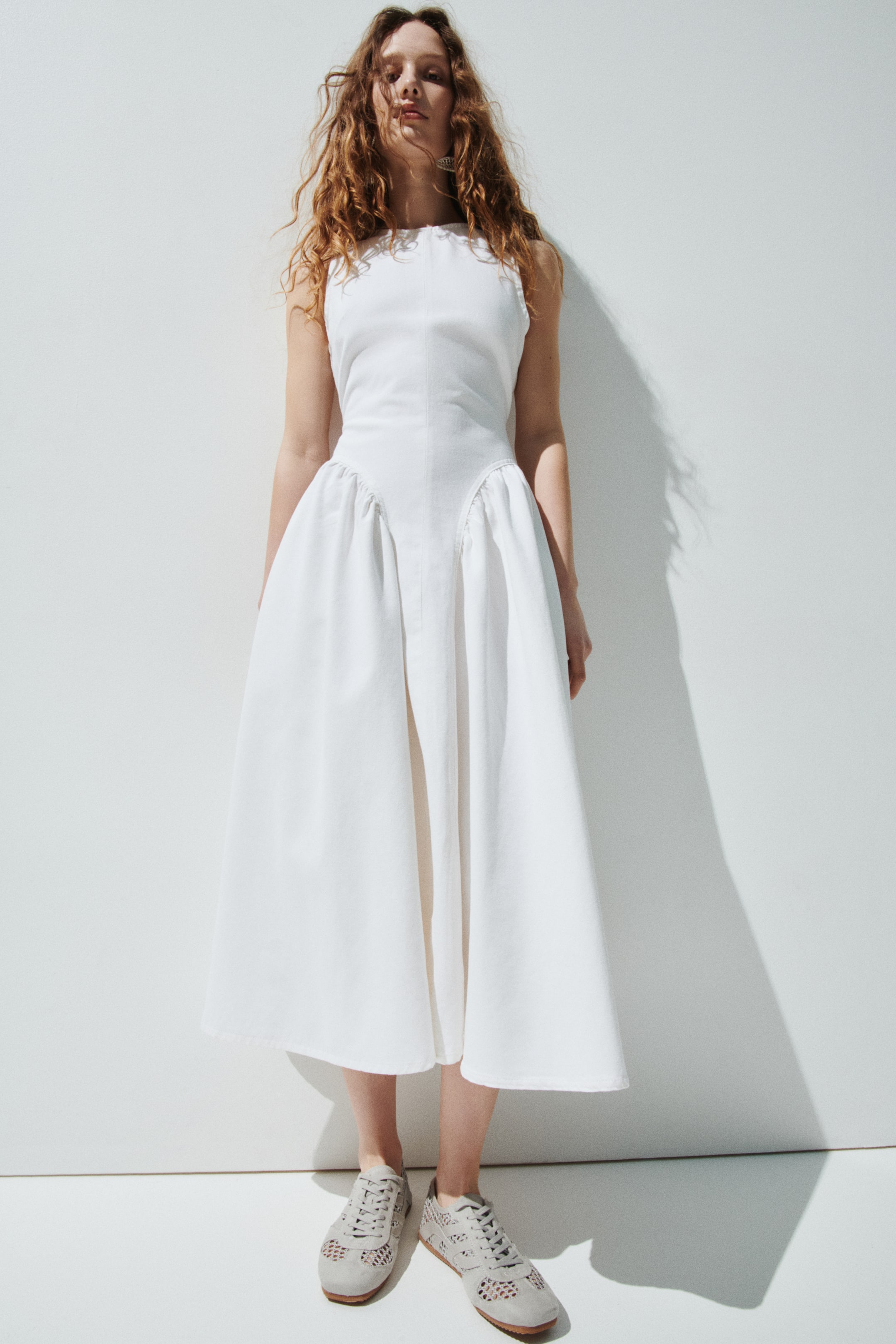 View larger image: A white sleeveless dress featuring a high round neckline and a fitted bodice with central vertical seams. The dress flares into a voluminous midi skirt, designed with prominent gathered side panels and a flat central front panel, extending to mid-calf.