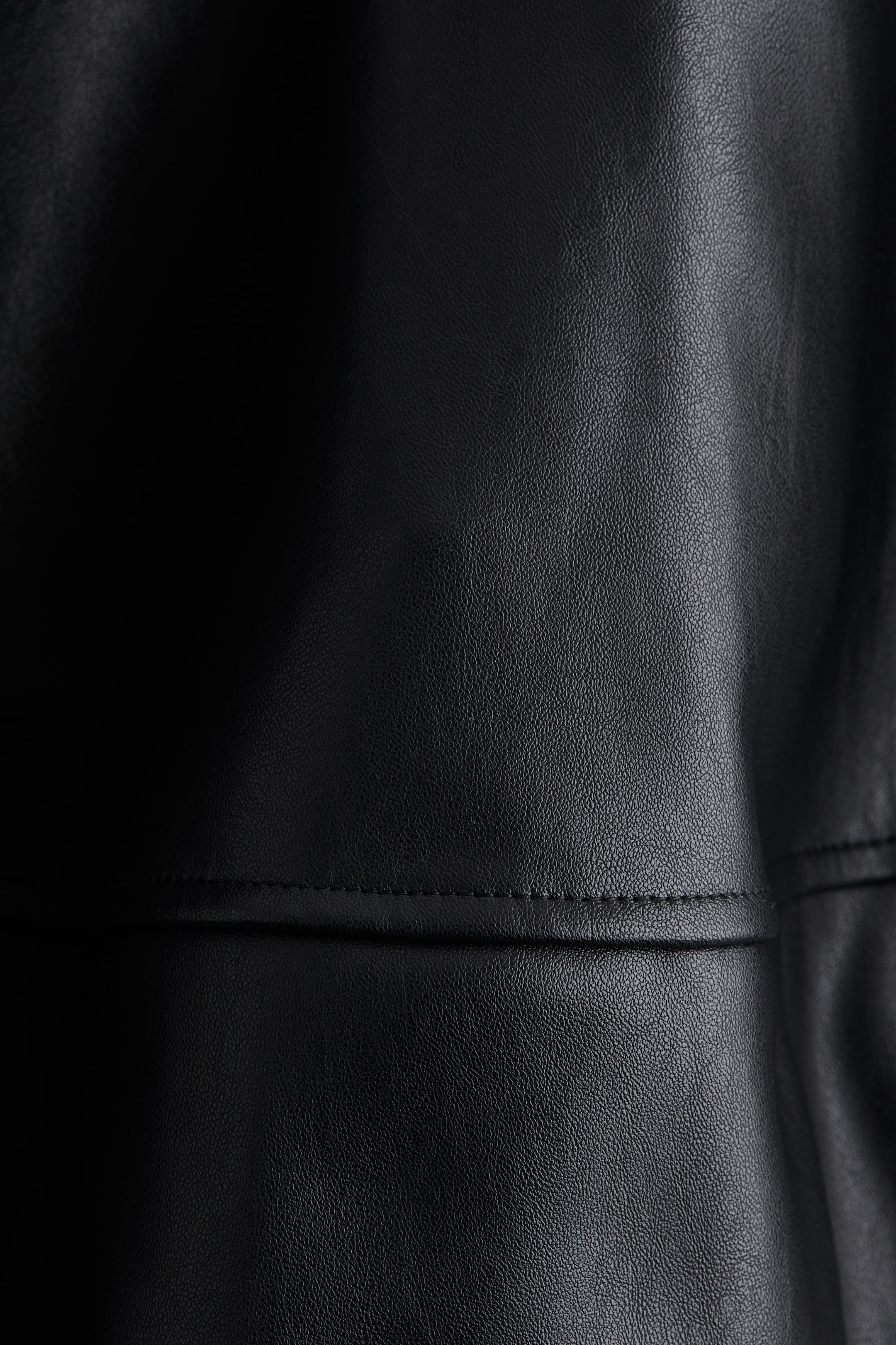 Vezi imaginea mai mare: Un material din piele neagră afișează o textură cu granulație fină. O cusătură orizontală cu cusături vizibile traversează suprafața sa, care prezintă, de asemenea, cute subtile.