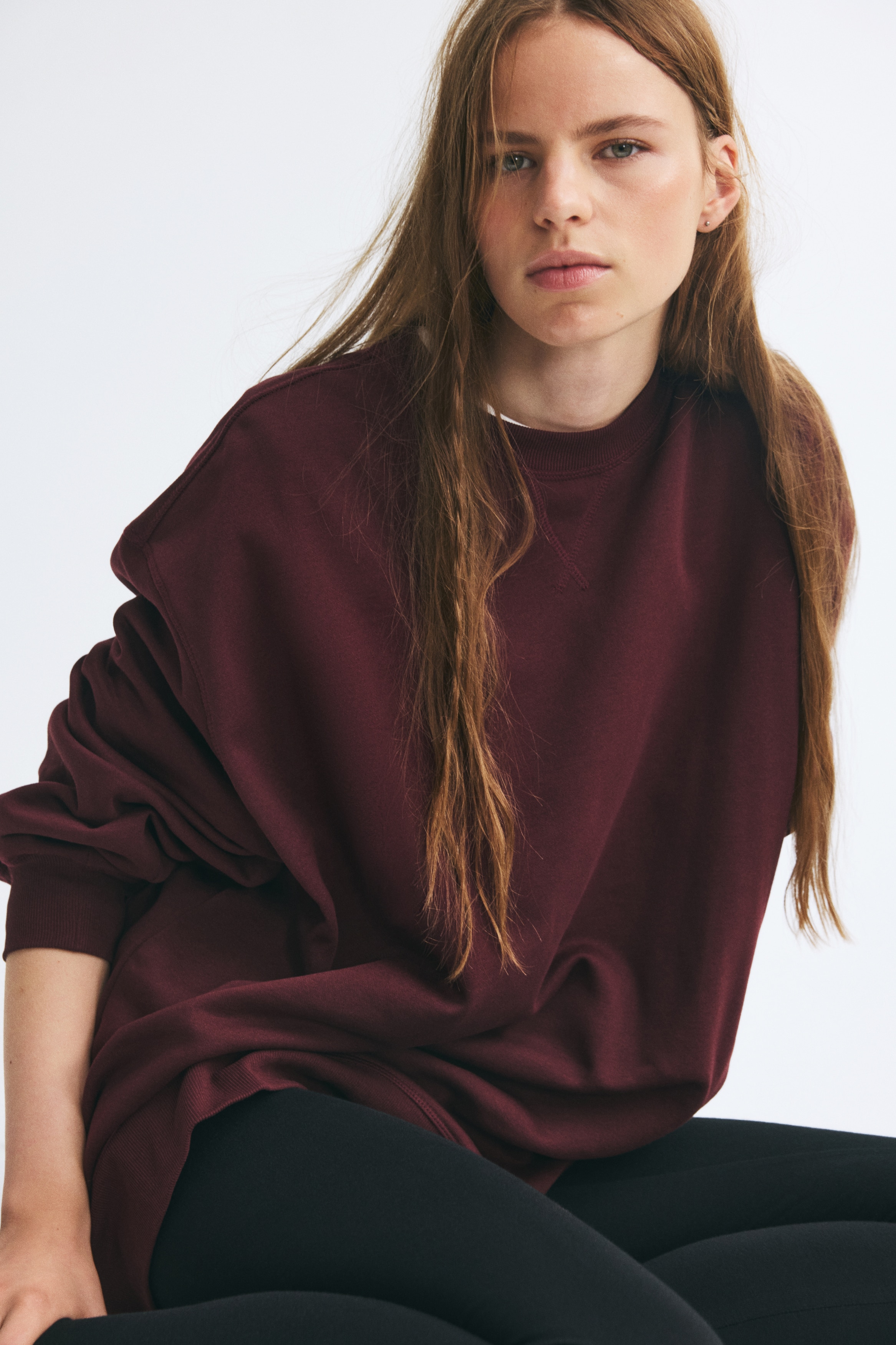 Ein oversized bordeauxfarbenes Sweatshirt hängt frontseitig und zeigt überschnittene Schultern, lange Ãrmel mit Rippbündchen sowie einen gerippten Rundhalsausschnitt mit gestepptem V-Detail. Das Kleidungsstück präsentiert eine entspannte, locker geschnittene Silhouette mit einem breiten gerippten Saum.