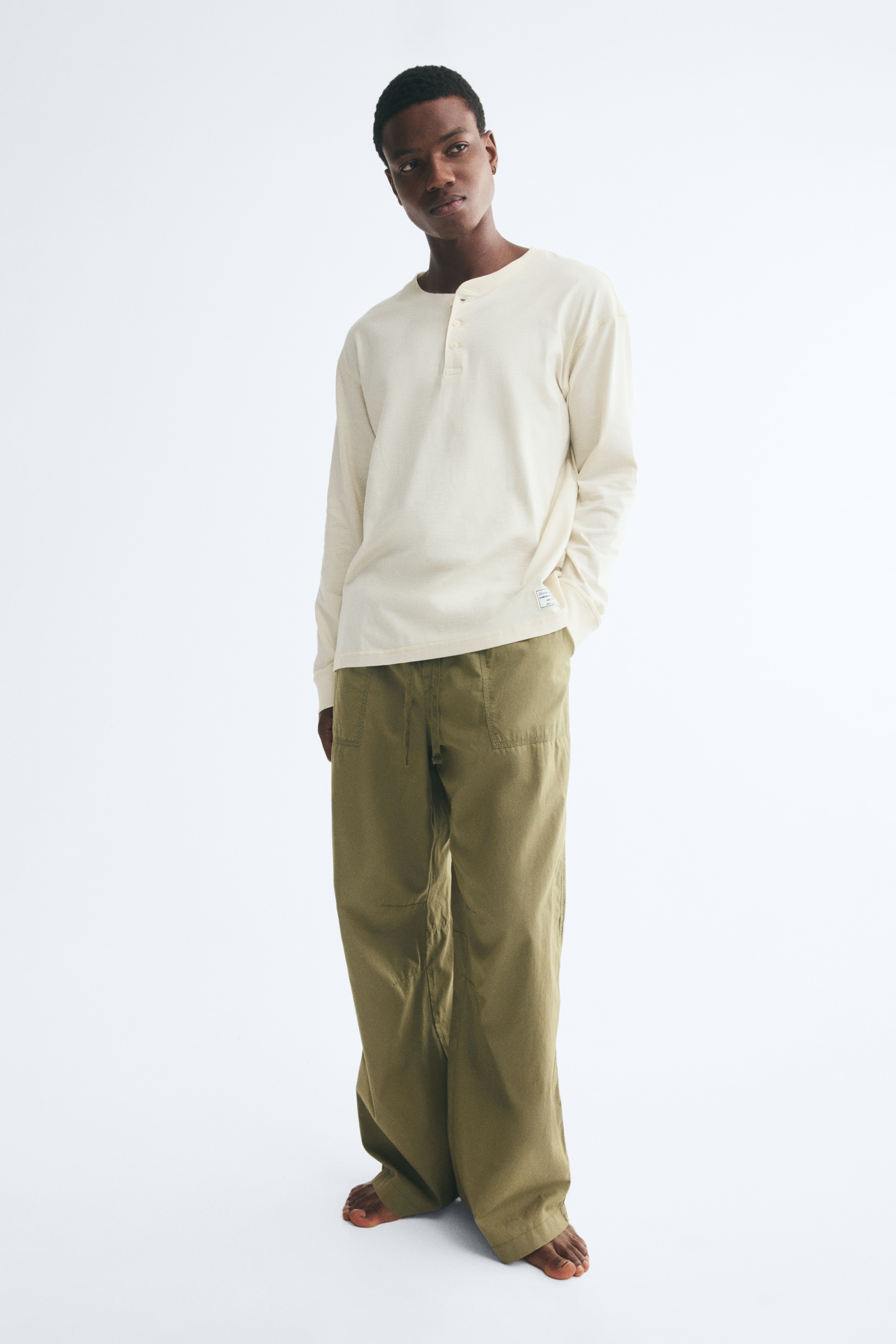 Una maglia Henley color avorio e pantaloni verde oliva sono presentati piegati e rivolti frontalmente come un set coordinato. La maglia presenta uno scollo rotondo a coste con abbottonatura a tre bottoni, mentre i pantaloni mostrano una vita elasticizzata con coulisse e una tasca laterale impuntata.