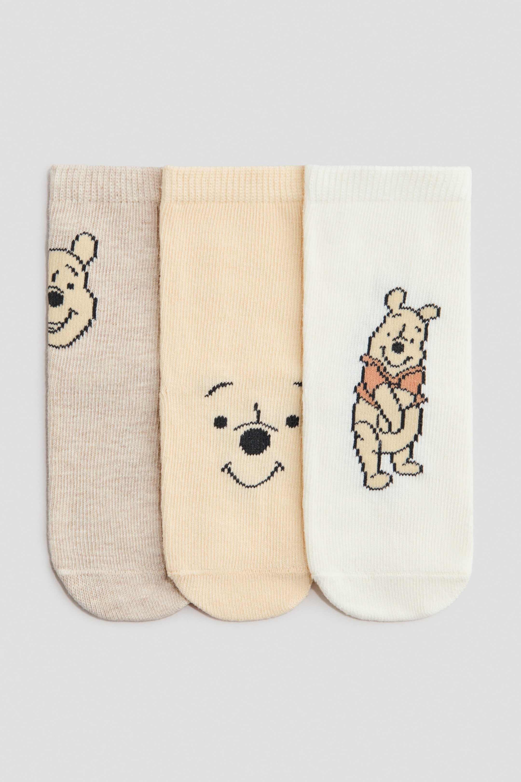 View larger image: Presenting a trio of fine-knit socks, each adorned with a charming Winnie the Pooh motif and featuring elasticated ribbed tops. The left sock, in a soft beige hue, showcases a partial face of Winnie the Pooh. The middle sock, in a light yellow shade, displays Pooh's full smiling face. Meanwhile, the right sock, in off-white, depicts a full-body image of Winnie the Pooh in his iconic red shirt.