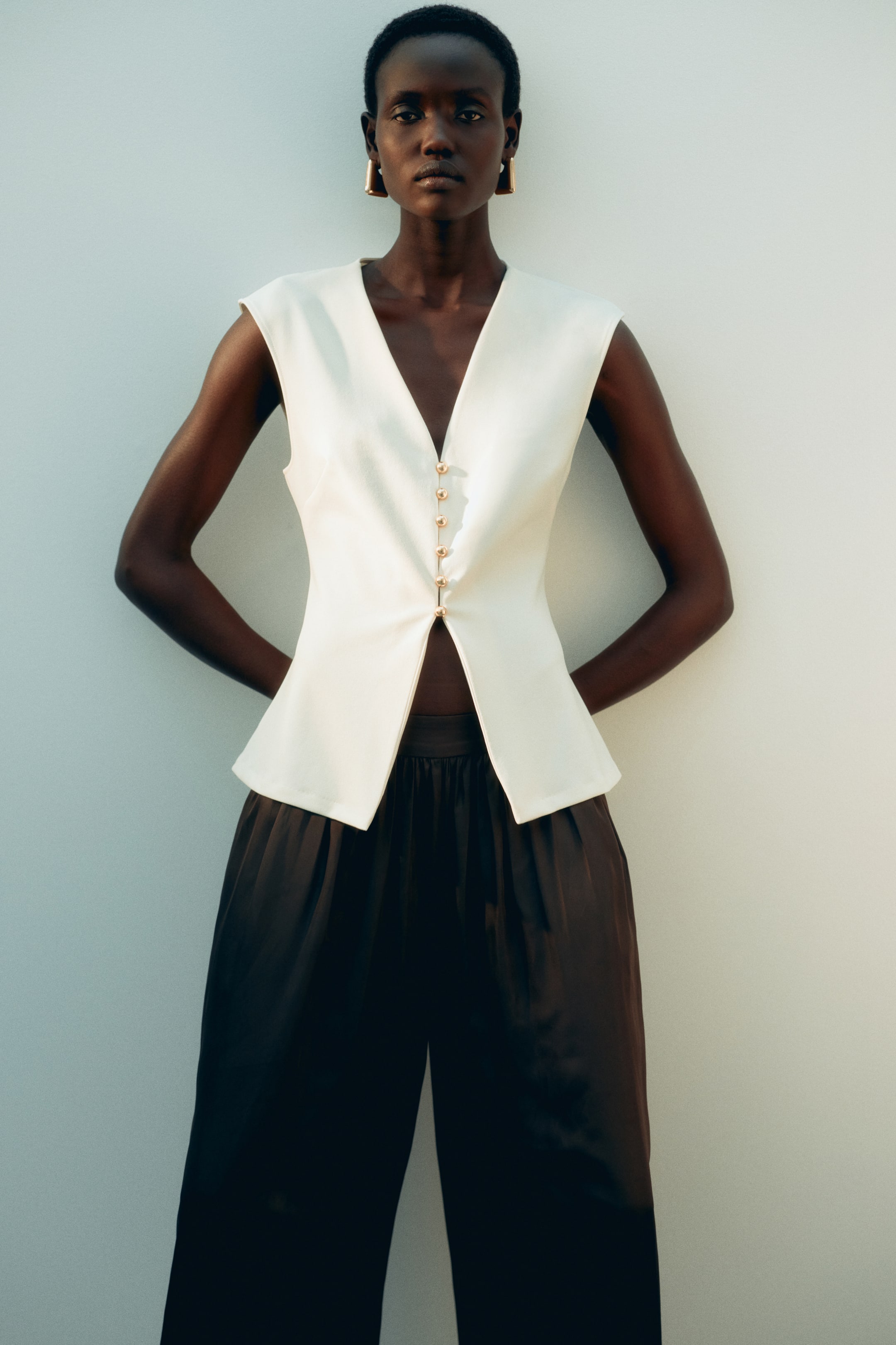 Xem ảnh lớn hơn: Mẫu mặc một chiếc áo gile ôm màu kem, cổ chữ V, với các cúc màu vàng bóng dạng vòm kéo dài đến eo. Áo có phần lai hơi loe và được phối bên ngoài quần ống rộng màu tối.