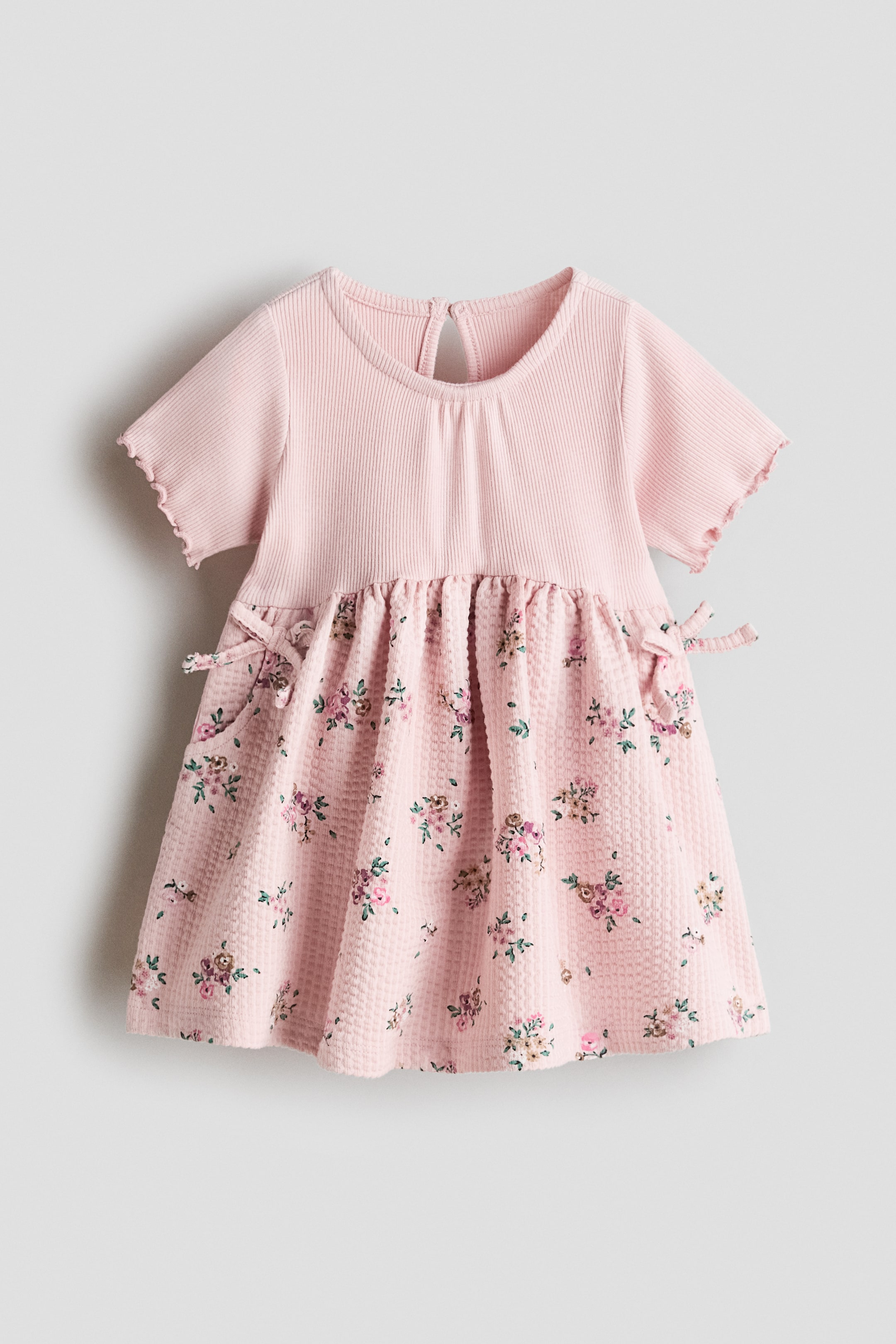 View larger image: This baby dress features a pink ribbed-knit bodice with short lettuce-edge sleeves and a round neckline, with a keyhole opening at the back. The gathered skirt is in pink waffle-knit fabric with a scattered floral print in pink, purple and green, and small decorative bows on either side of the waist.
