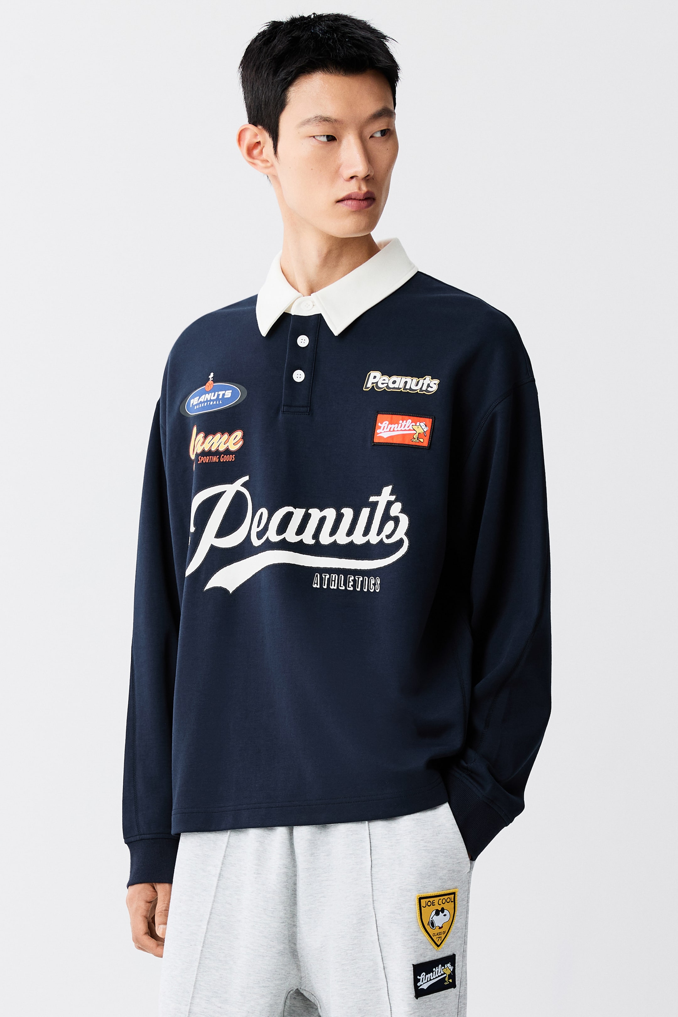 View larger image: A navy blue, loose-fit rugby shirt featuring a white collar, a button placket, and printed Peanuts-themed graphics is being worn by the model. The shirt has dropped shoulders and long sleeves with ribbed cuffs, and is paired with light grey sweatpants featuring decorative patches.