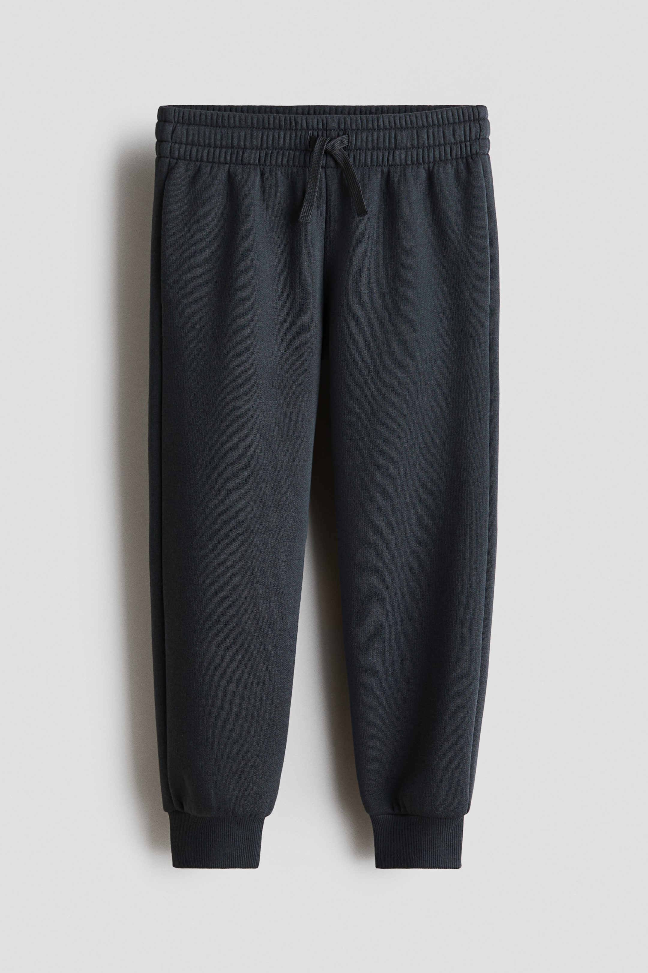 View larger image: Dark gray joggers shown from the front, featuring an elasticated waistband with a tied drawstring. Side pockets are visible along the seams, and the ankles are finished with ribbed cuffs.