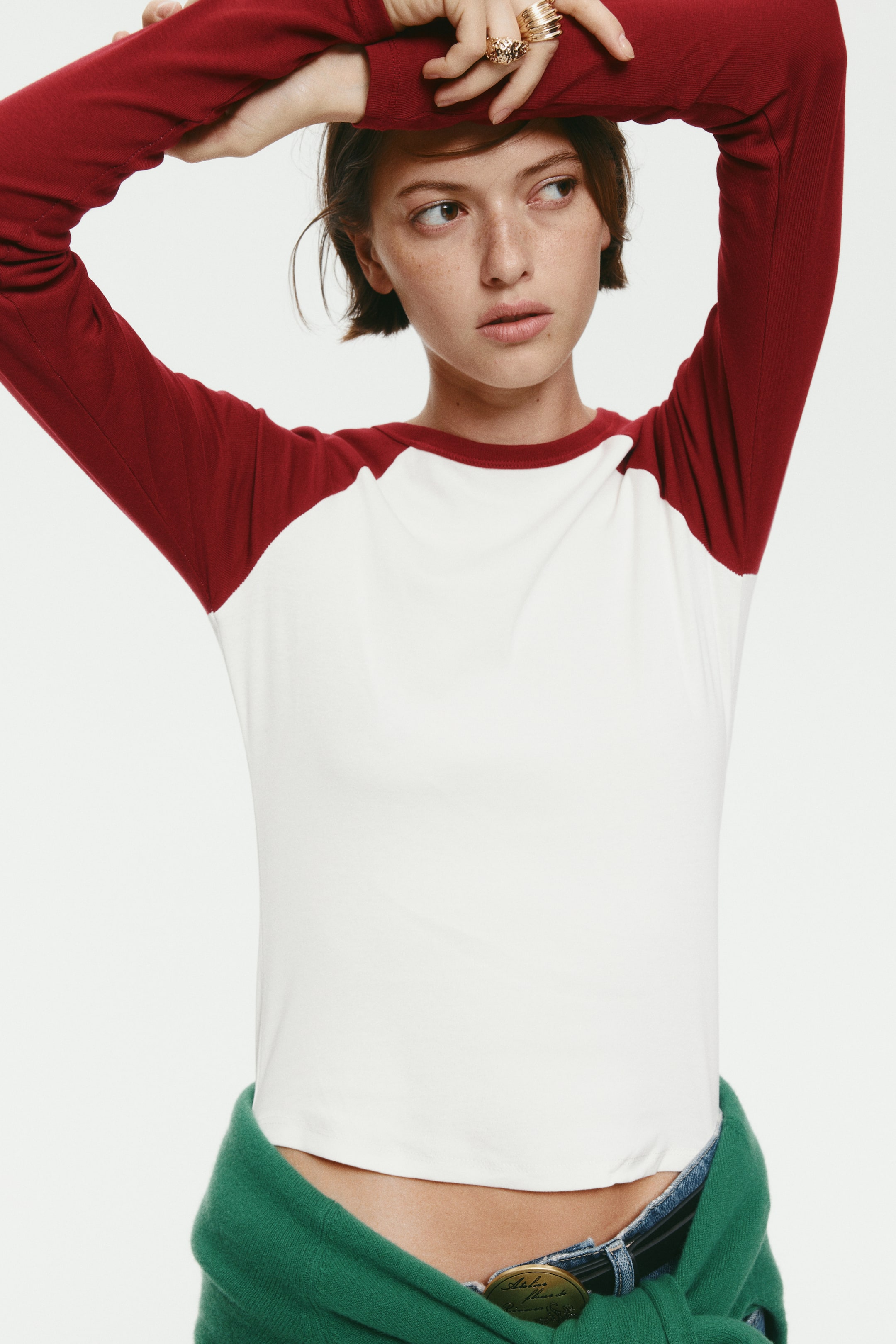 Se større billede: En tætsiddende raglan-top med hvidt hovedstykke, rød rund halsudskæring og lange røde ærmer bæres af modellen, som poserer med armene løftet. Toppen styles med blå jeans og en grøn sweater bundet om талjen.