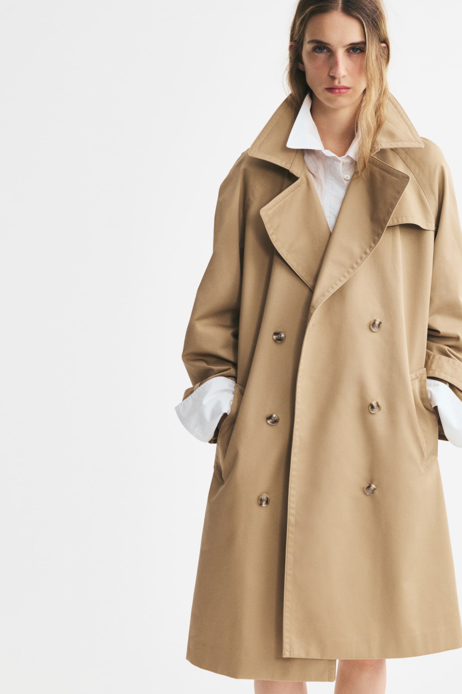 A tan double-breasted trench coat showcases notch lapels, a storm flap, and a matching fabric belt tied at the waist. It features adjustable cuff straps on the long sleeves and brown buttons down the front. - 1
