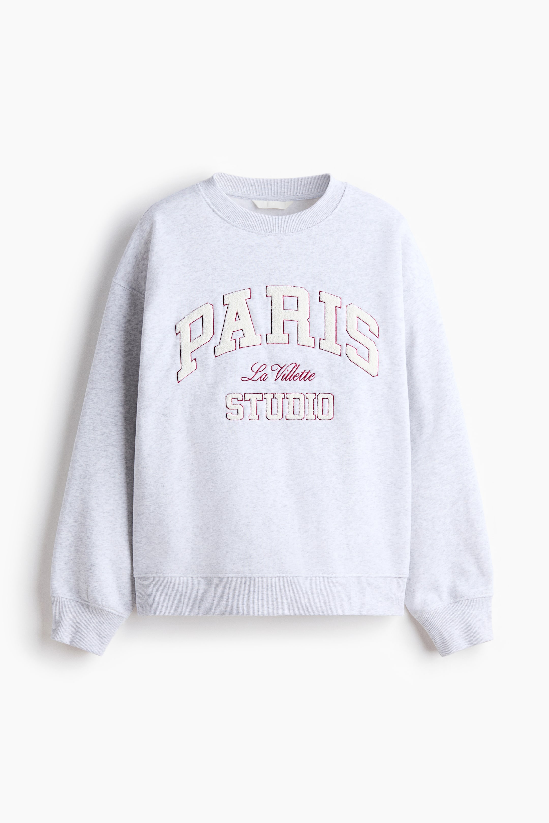 View larger image: The oversized light gray heathered sweatshirt is showcased from the front, featuring a round, ribbed neckline, dropped shoulders, and long sleeves with ribbed cuffs. The front displays a text motif with 'PARIS' in arched, cream-filled letters outlined in red, 'La Villette' in red script, and 'STUDIO' in cream-filled letters outlined in red, above a ribbed hem.