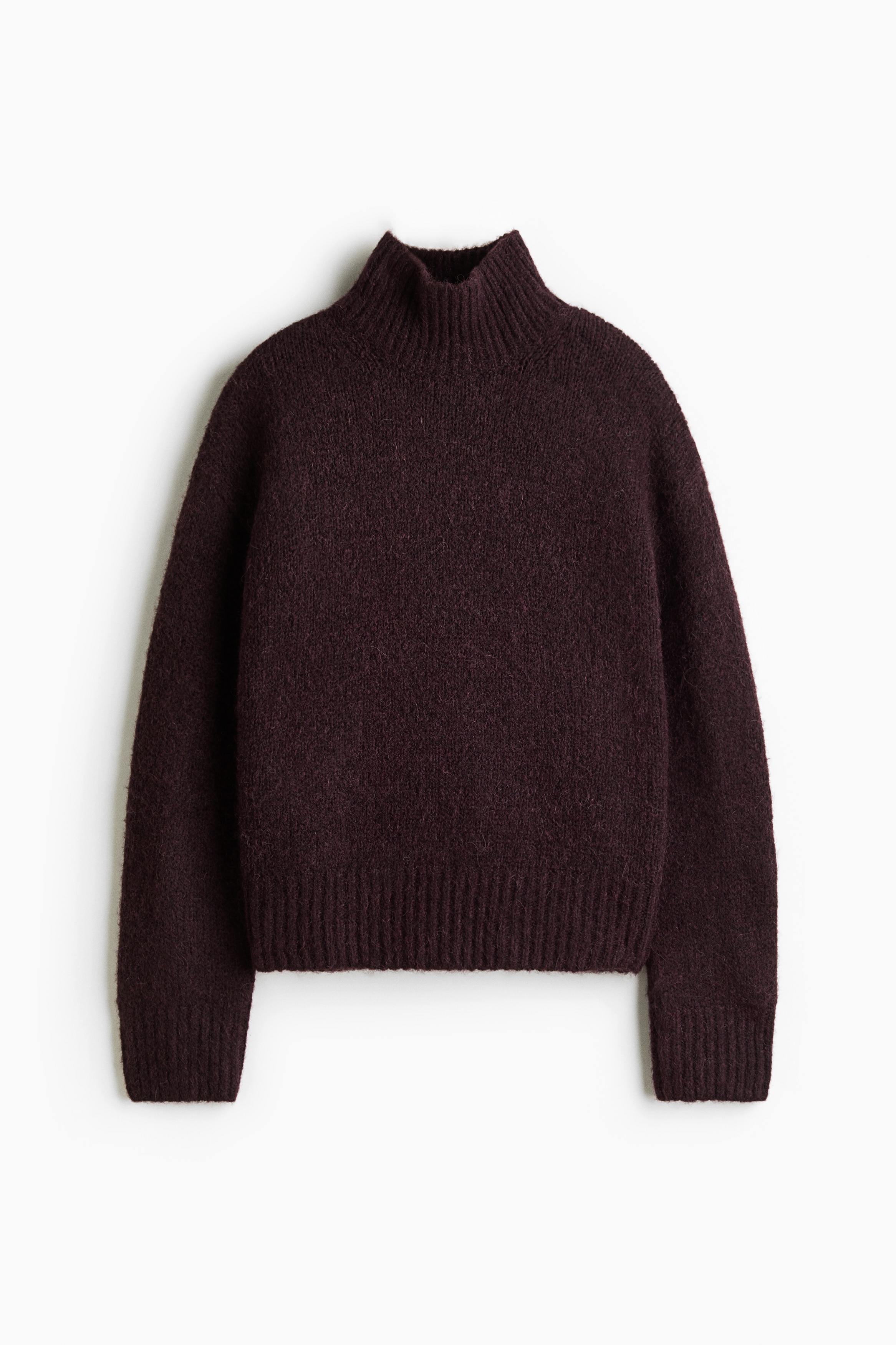 A dark burgundy knitted sweater with a brushed finish is being worn by the model, featuring a high ribbed neck, dropped shoulders, and ribbed cuffs. The sweater is styled with a pair of wide-leg blue jeans and a black belt.