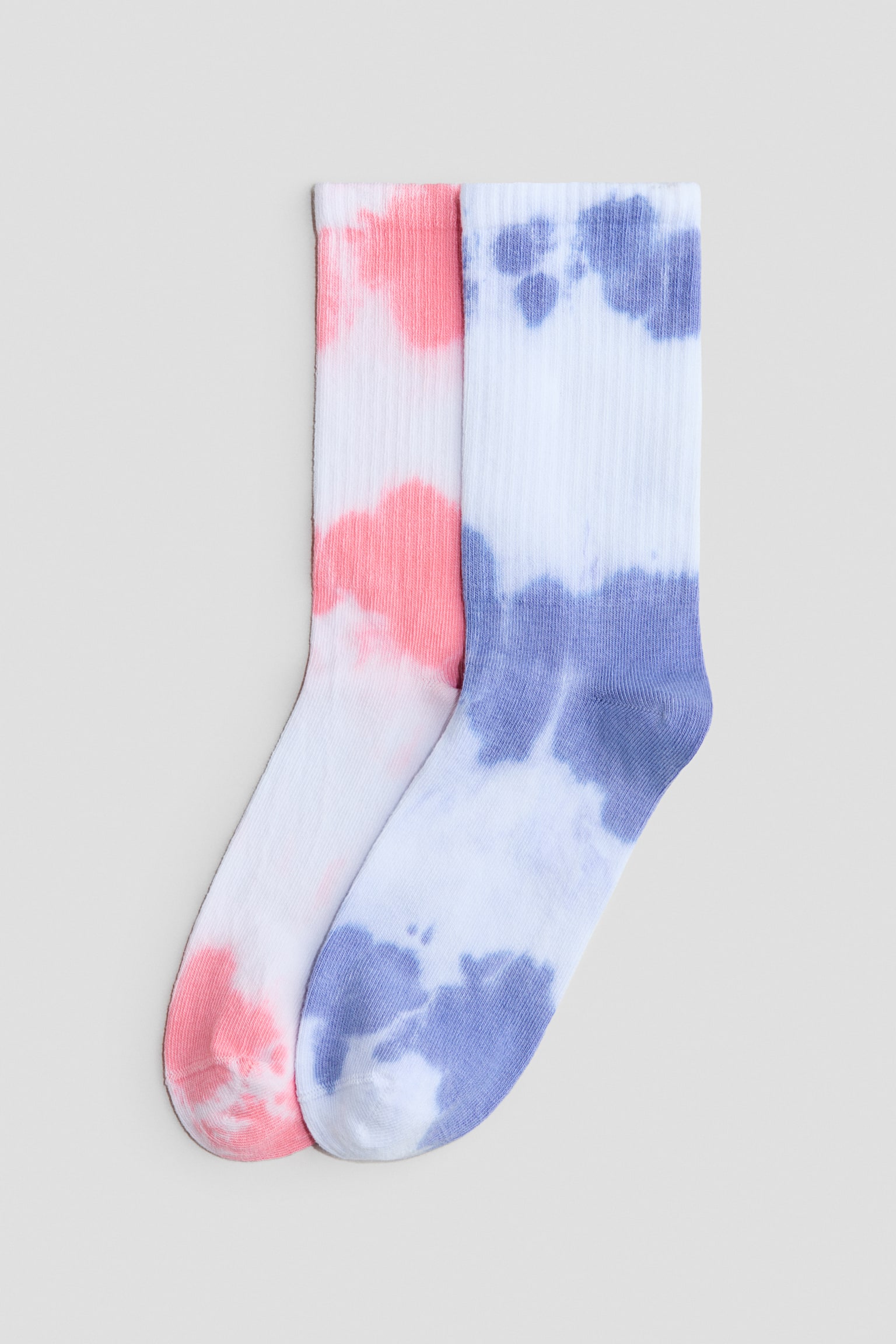 Een paar witte geribde crewsokken wordt van voren getoond met een tie-dye patroon, waarbij de ene sok koraalroze vlekken heeft en de andere blauw-violette vlekken. De sokken liggen plat en overlappen lichtjes, waardoor de geribde textuur langs de schacht en de gladde brei aan de voet worden geaccentueerd.