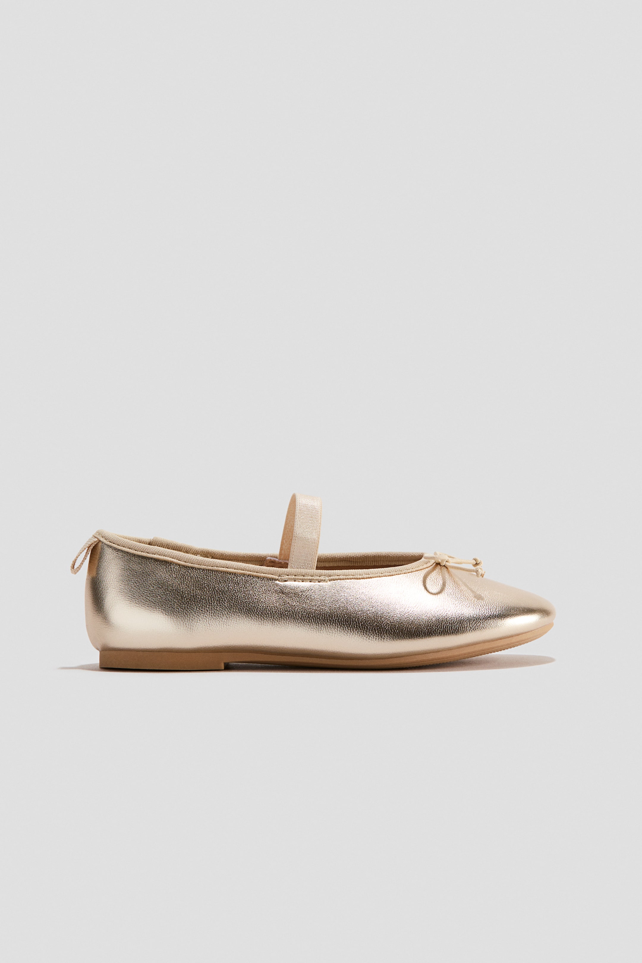 View larger image: A gold ballet pump is resting on its side, angled slightly towards the front. It features a rounded toe with a decorative bow, an elasticated foot strap across the instep, grosgrain trim around the opening, and a small loop at the back.