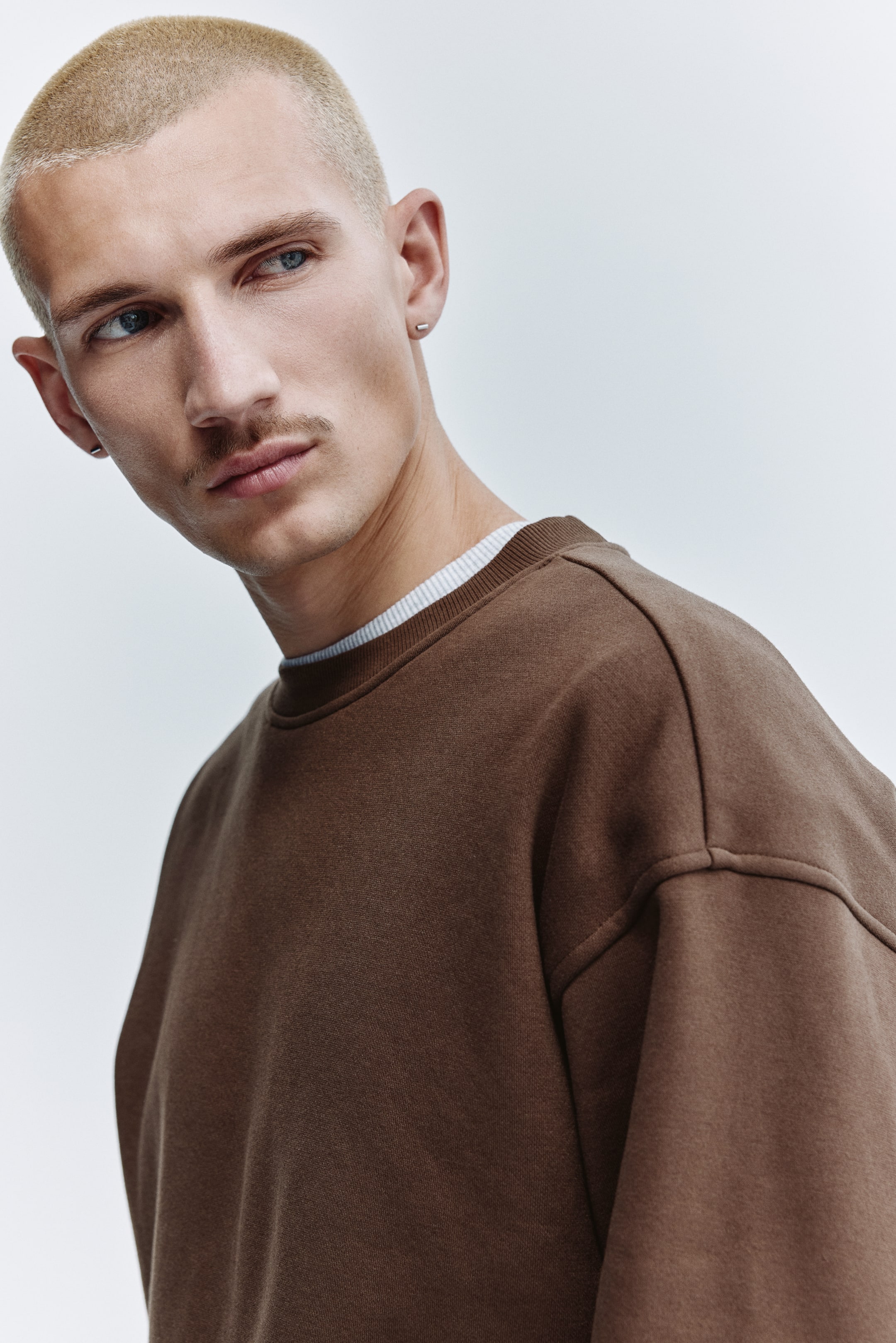 View larger image: The brown crewneck sweatshirt features a ribbed neckline and visible shoulder seams, shown in a relaxed fit from a front-side angle. A light-colored, possibly striped, undershirt peeks out from beneath the collar.