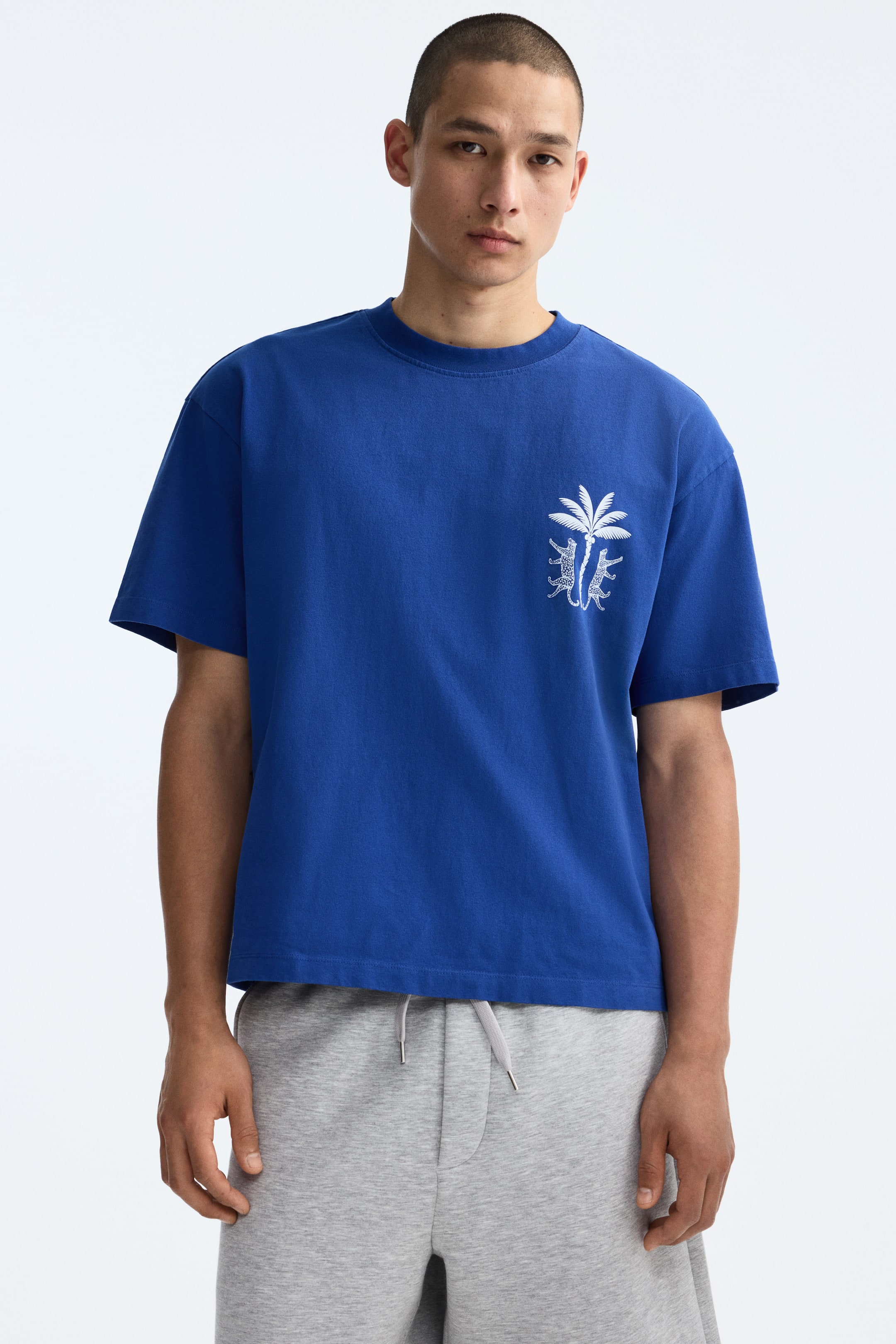 View larger image: The model is wearing a blue boxy-style T-shirt made from cotton jersey. The T-shirt features a white print motif of a palm tree and two leopards on the front, along with a round rib-trimmed neckline, dropped shoulders, and a straight-cut hem. The model is also paired with gray sweat shorts.