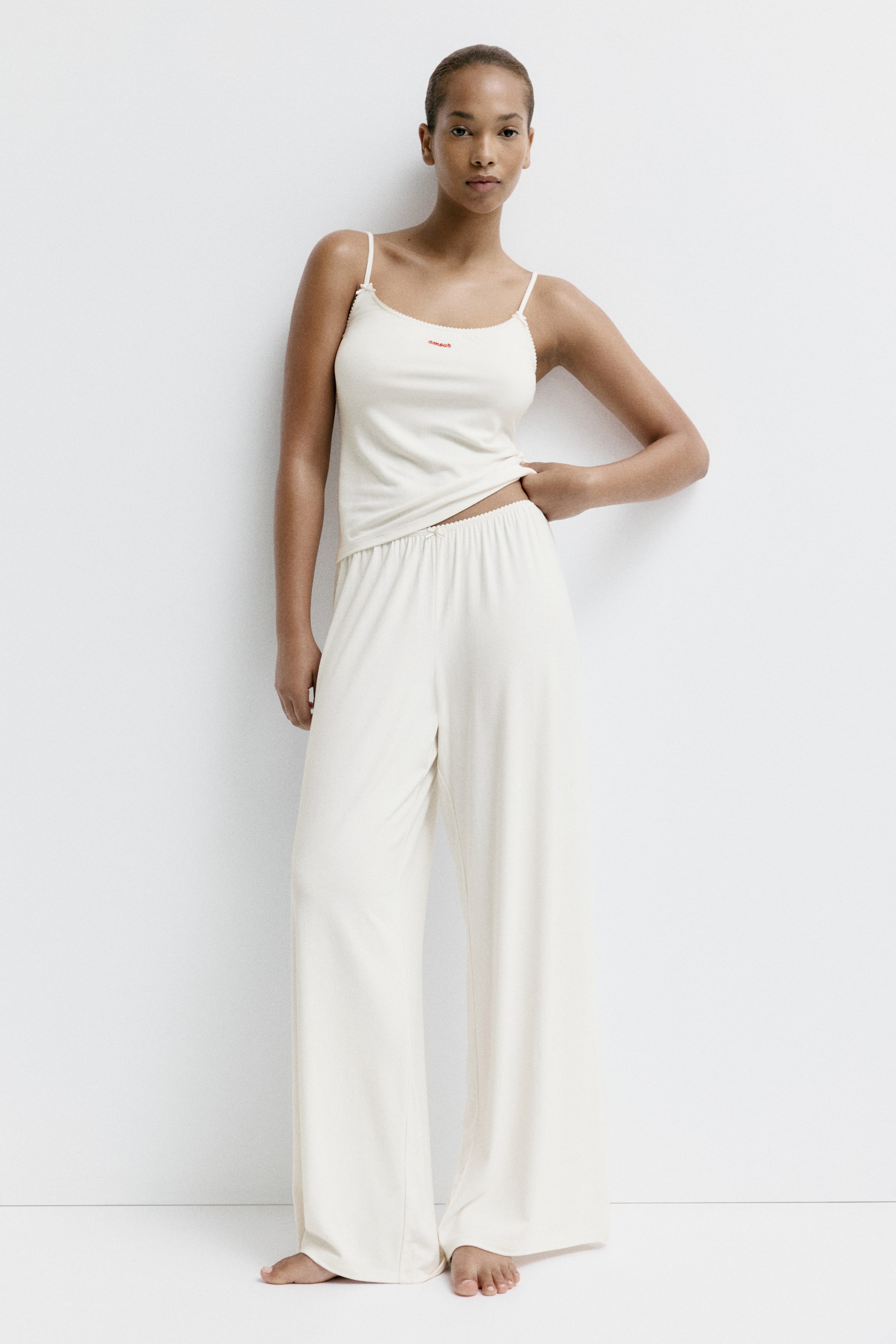 View larger image: A cream two-piece loungewear set features a camisole top with thin spaghetti straps, scalloped trim, and red embroidered text on the chest. The matching wide-leg pants have an elasticated waistband and a loose, flowing fit.