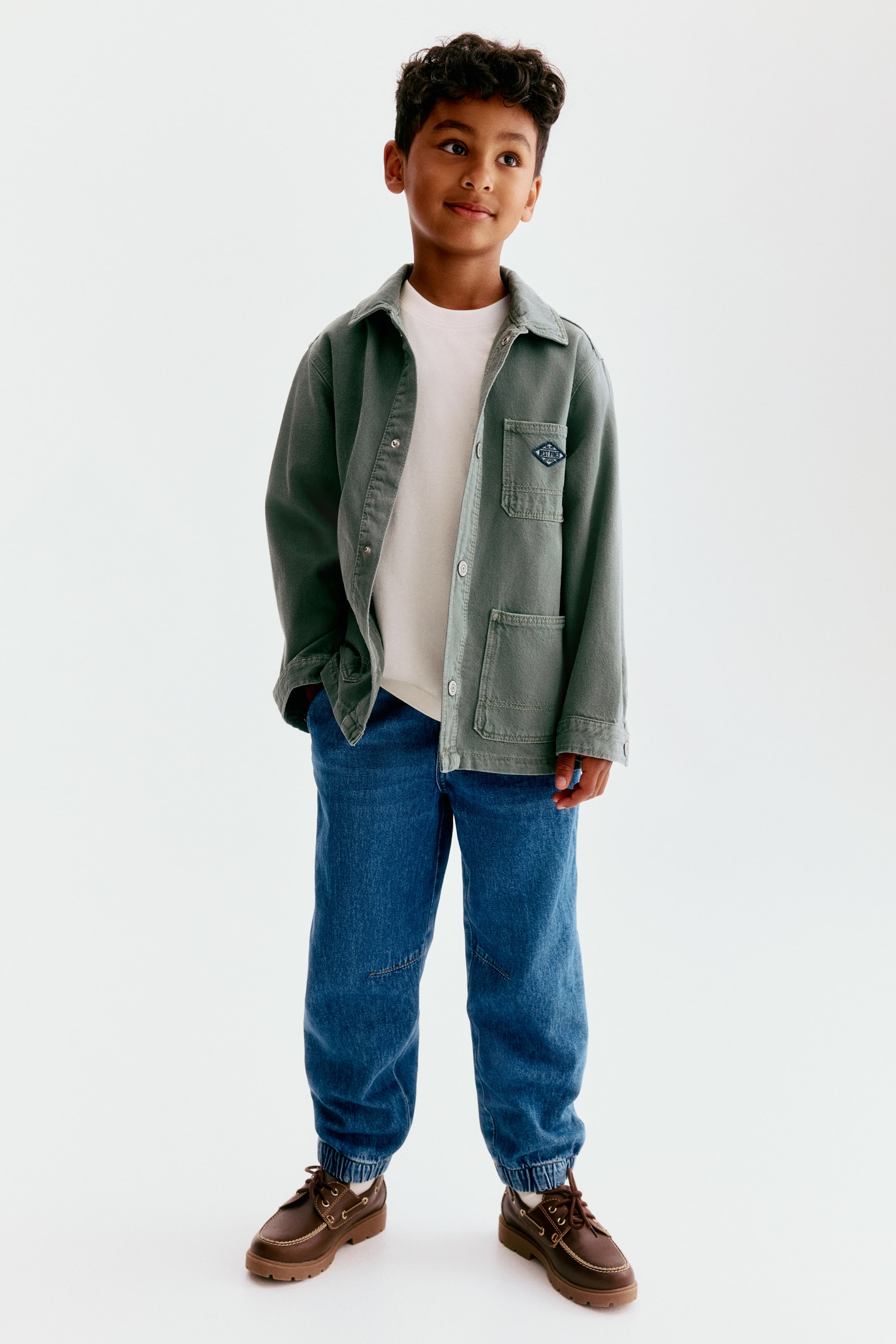 Größeres Bild anzeigen: Oversized-Hemdjacke aus Baumwolle. Diese verblasste grüne Oversized-Hemdjacke aus Baumwolle ist von vorne zu sehen und verfügt über einen spitzen Kragen, Knopfverschlüsse vorne, zwei große aufgesetzte Taschen unten und eine aufgesetzte Brusttasche mit Rauten-Emblem. Sie wird offen über einem weißen T-Shirt getragen.