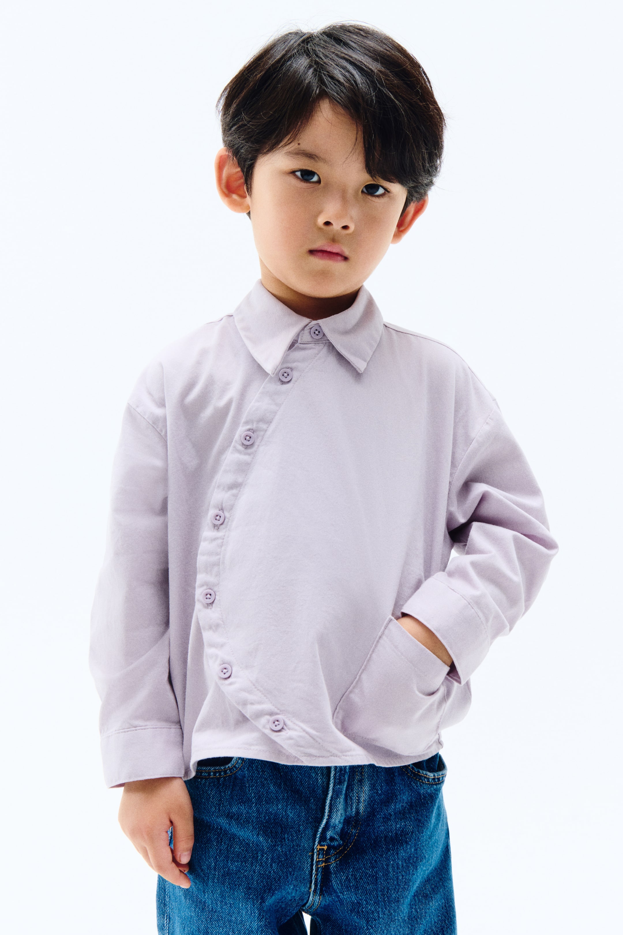 拡大画像を表示: ライトパープルの長袖シャツは、右肩から左ヒップにかけてカーブした非対称ボタンのプラケットと尖った襟が特徴です。ボタン付きカフスの長袖を備えており、手がサイドポケットに入れられ、優雅なドレープを作り出しています。