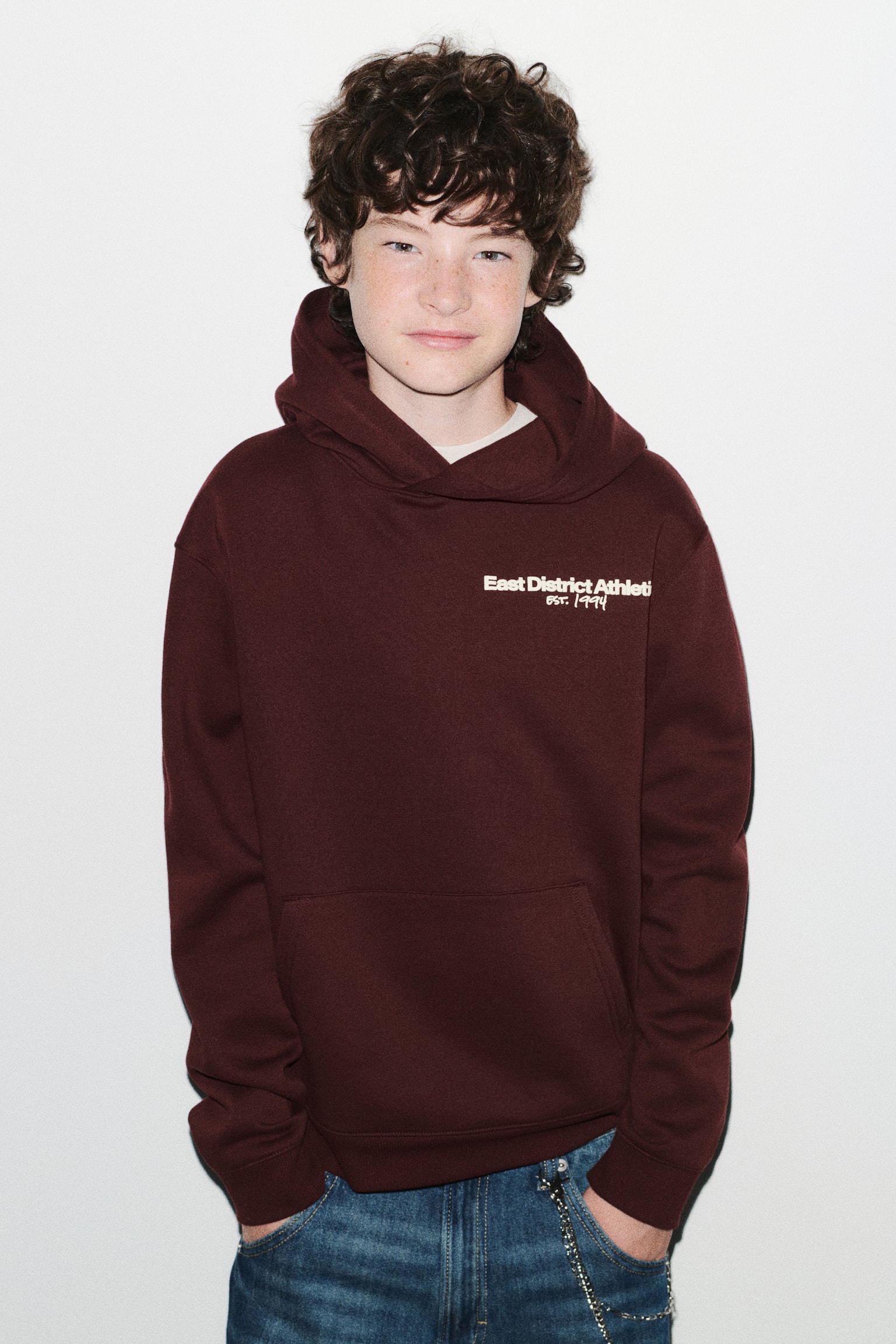 A front-facing view shows a dark burgundy hoodie featuring a small white text print on the left chest that reads "East District Athletic Est. 1994". The hoodie is designed with a wrap-front hood, dropped shoulders, a kangaroo pocket, and ribbed cuffs and hem, and is shown styled with blue jeans. - 1