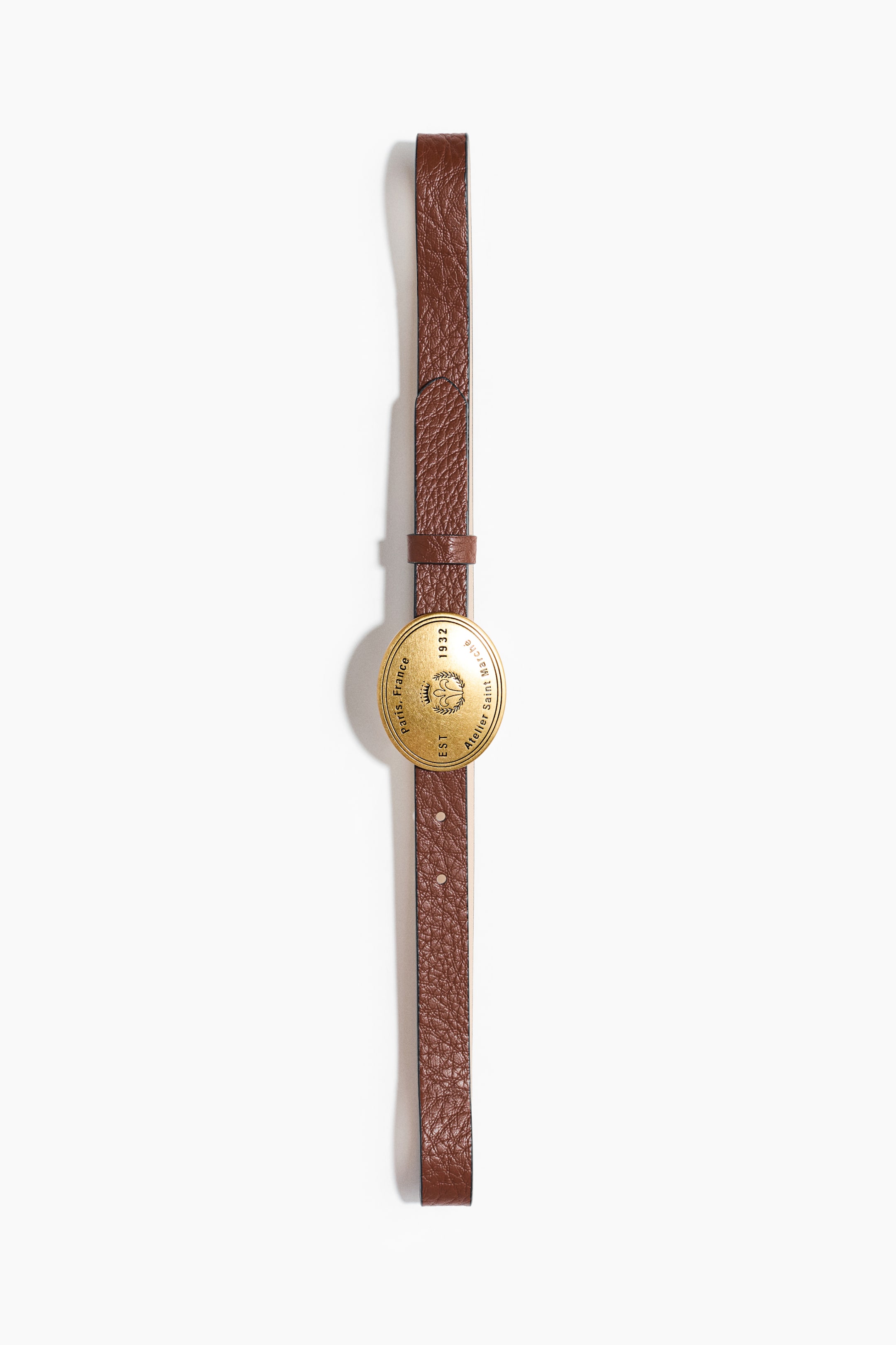 View larger image: The belt is a slim brown leather strap with a textured finish. Centrally placed on the strap is a round, gold-toned metallic emblem showcasing text such as 'Atelier Saint Marche', 'EST 1932', and 'paris, France' encircling a crown motif.