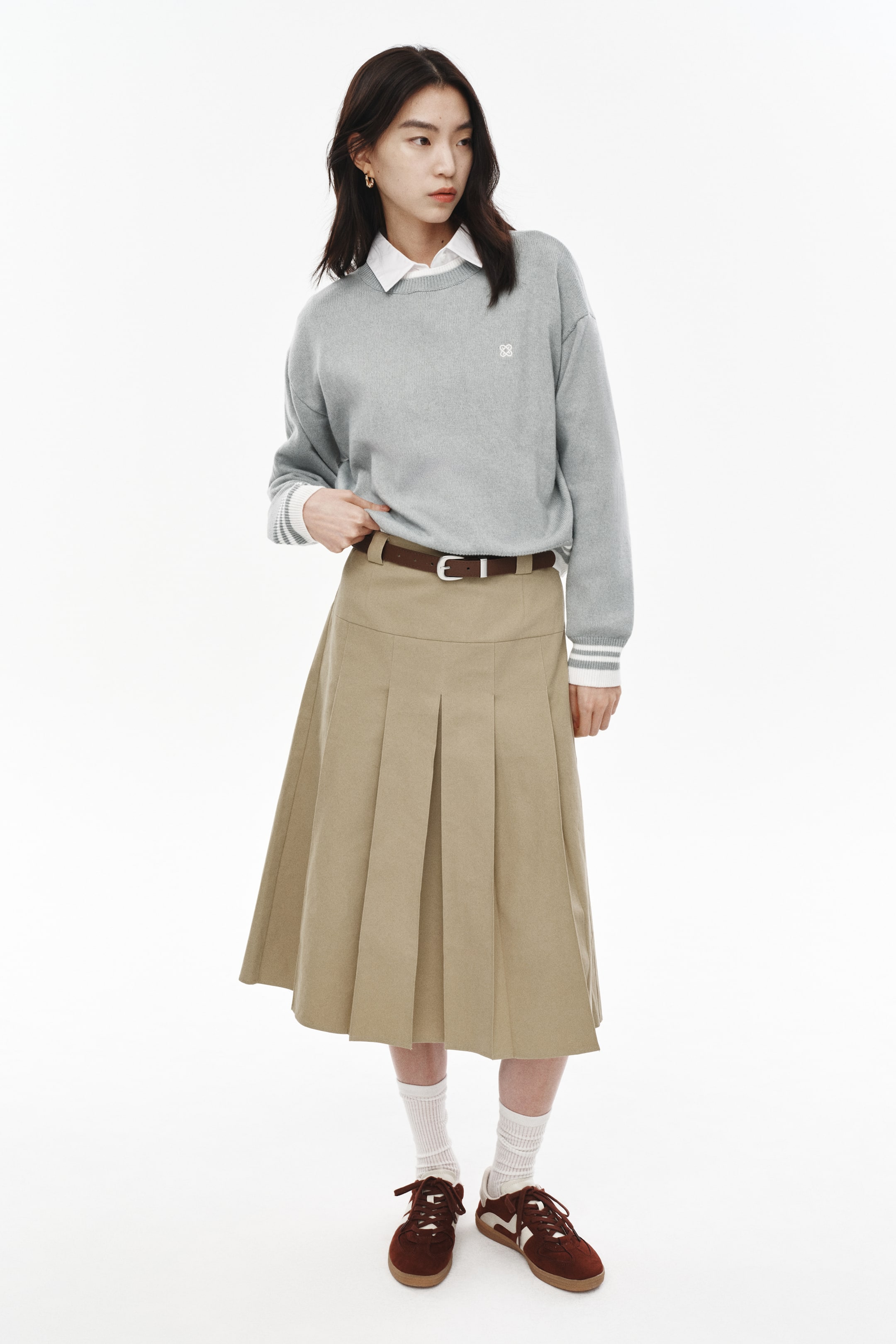 拡大画像を表示: ライトグレーのクルーネックセーターは、左胸に白い刺繍のロゴが施され、白いストライプの入ったリブ付きカフスが特徴です。このセーターは、白い襟付きシャツの上に着用されています。シルバーバックル付きのブラウンベルトがカーキ色のプリーツ入りミディスカートを締めており、白いリブ付きソックスと、白いアクセントとガムソールが特徴のダークブラウンのスエードスニーカーと組み合わせられています。