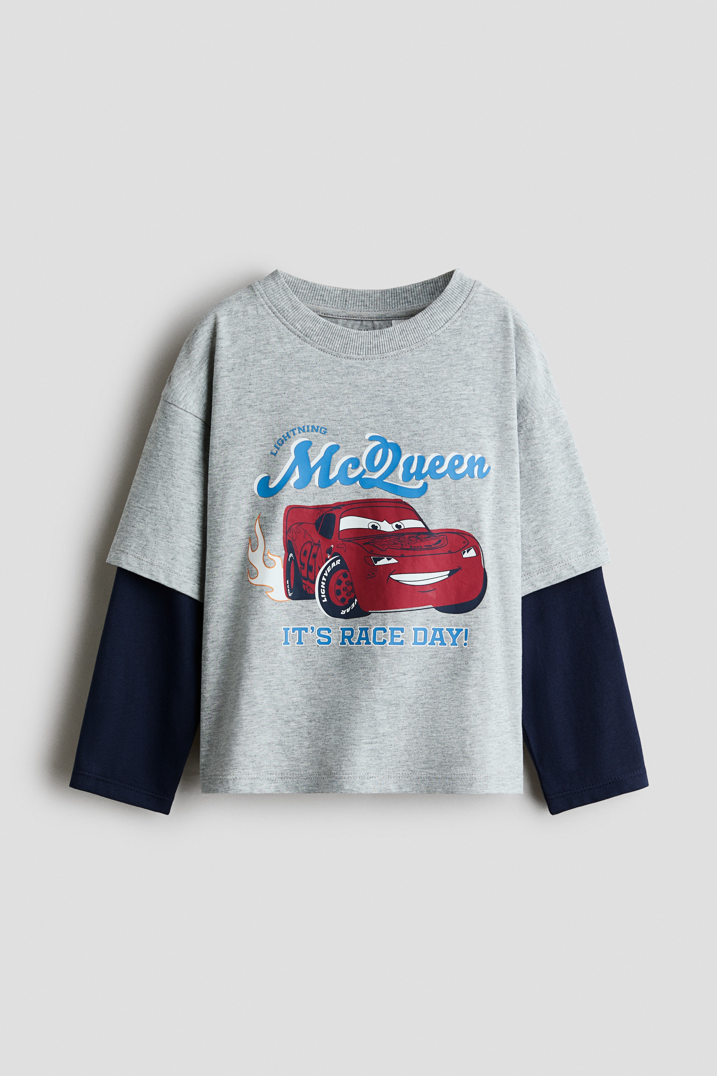 A light grey cotton jersey T-shirt, shown from the front, features a layered design with short sleeves over sewn-in navy blue long sleeves. The T-shirt has a round, rib-trimmed neckline, dropped shoulders, and a printed graphic of the red cartoon car Lightning McQueen with blue text reading âLightning McQueenâ and âITâS RACE DAY!â.