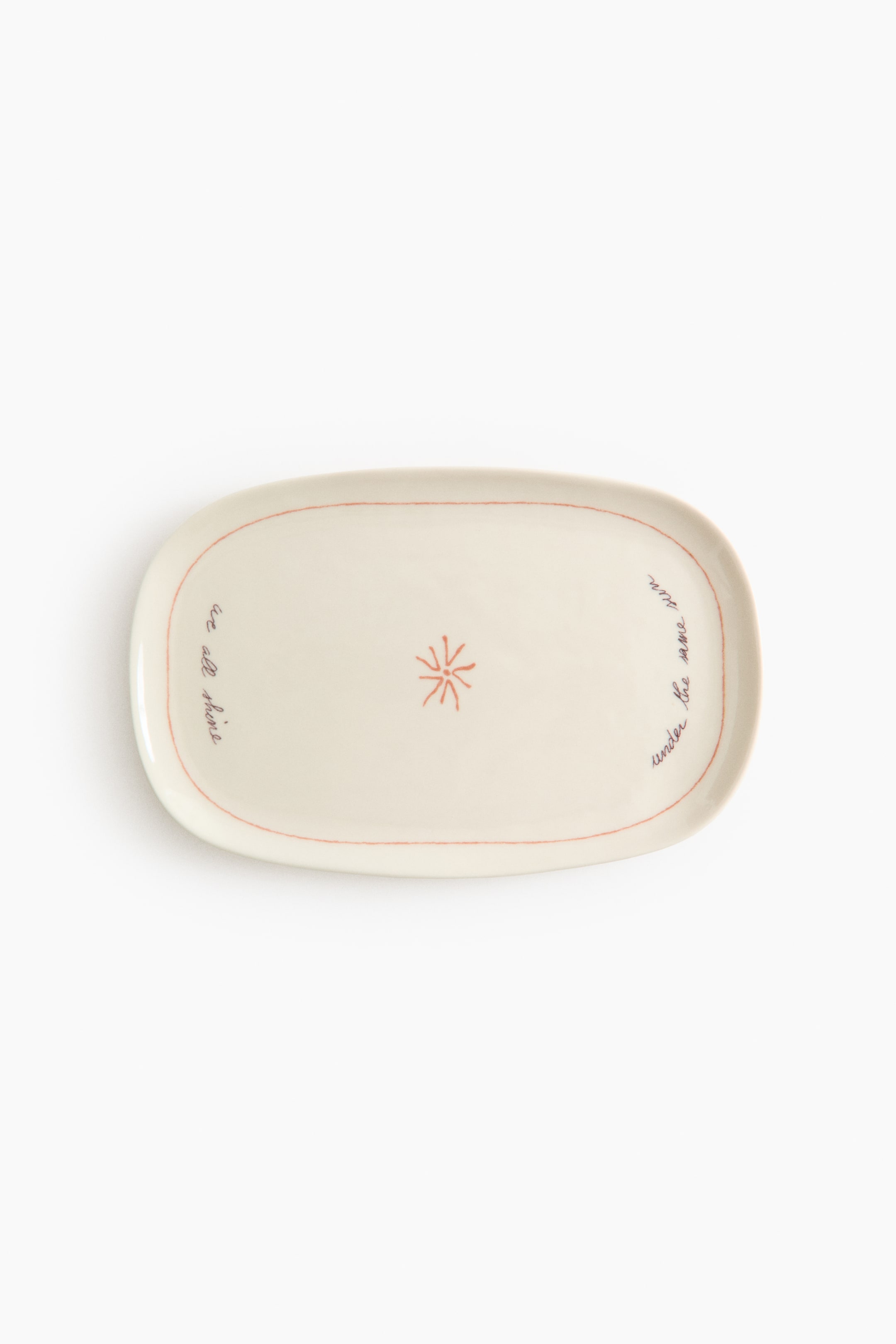 Voir une image plus grande: Un plateau de service ovale en céramique blanc cassé présente une ligne rougeâtre-orange qui trace son bord intérieur. Le plateau arbore un motif central en étoile et un texte manuscrit indiquant "nous brillons tous" à gauche et "sous le même soleil" à droite.
