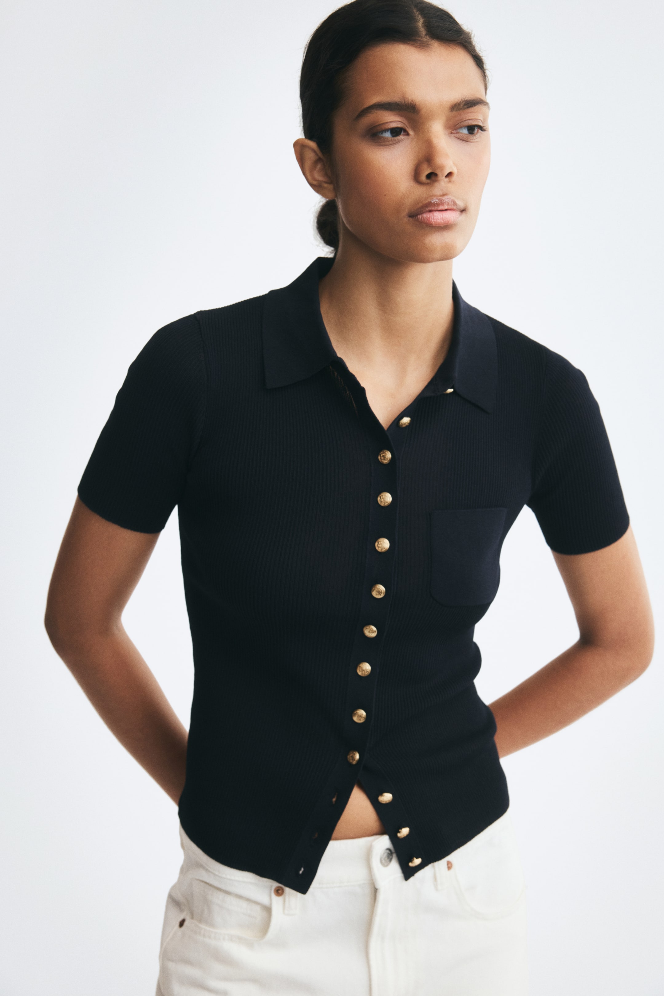 View larger image: A black, short-sleeved cardigan in a fine rib knit is worn by the model, featuring a collar, a patch pocket on the chest, and embossed gold-tone buttons down the front. The cardigan is styled with white trousers, with the bottom buttons left unfastened at the waist.