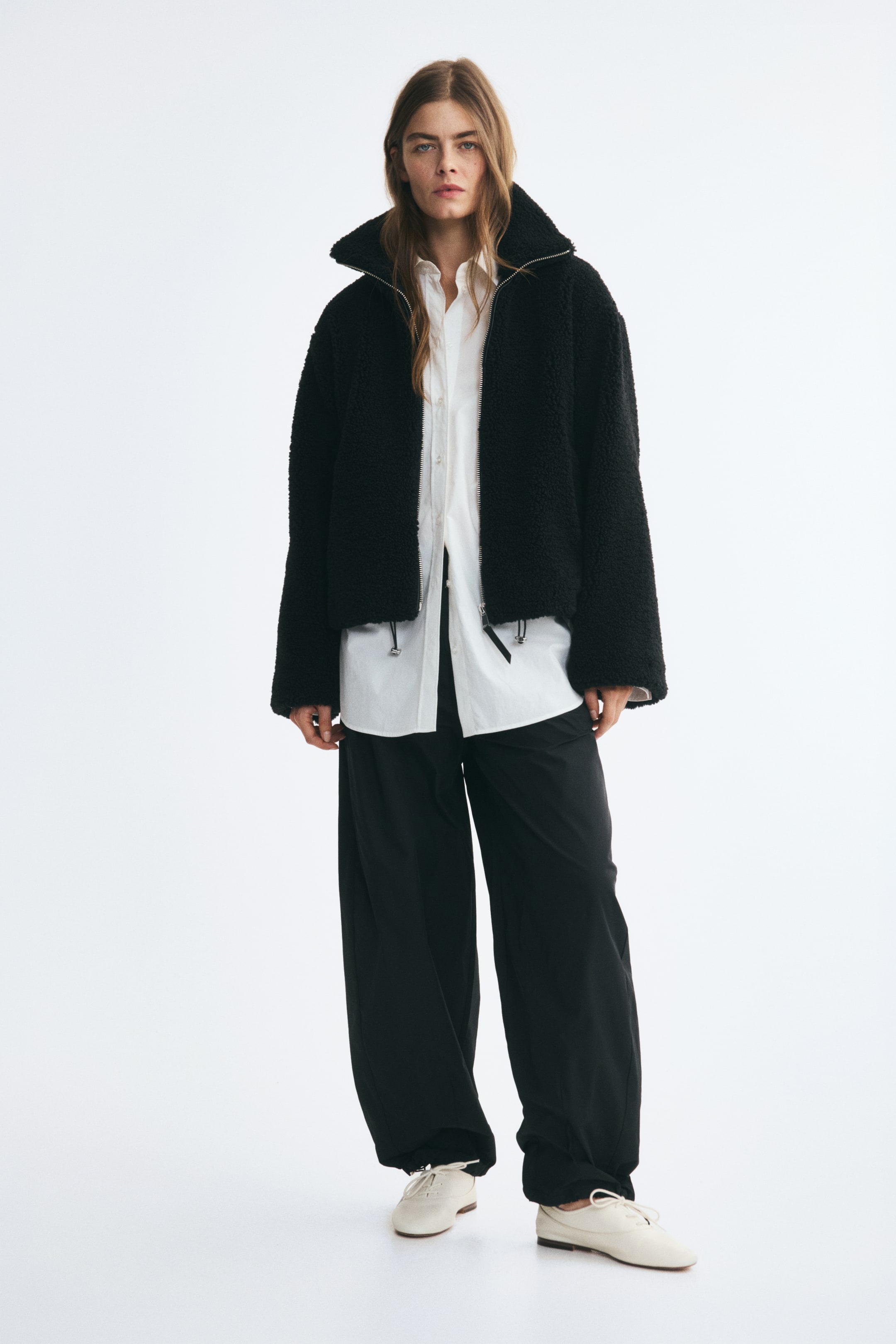 Größeres Bild ansehen: Eine schwarze Teddyjacke mit Stehkragen, überschnittenen Schultern und Tunnelzug am Saum wird vom Model offen getragen. Die Jacke ist über einem langen weißen Hemd mit Kragen getragen und mit einer schwarzen Weitbein-Hose kombiniert.