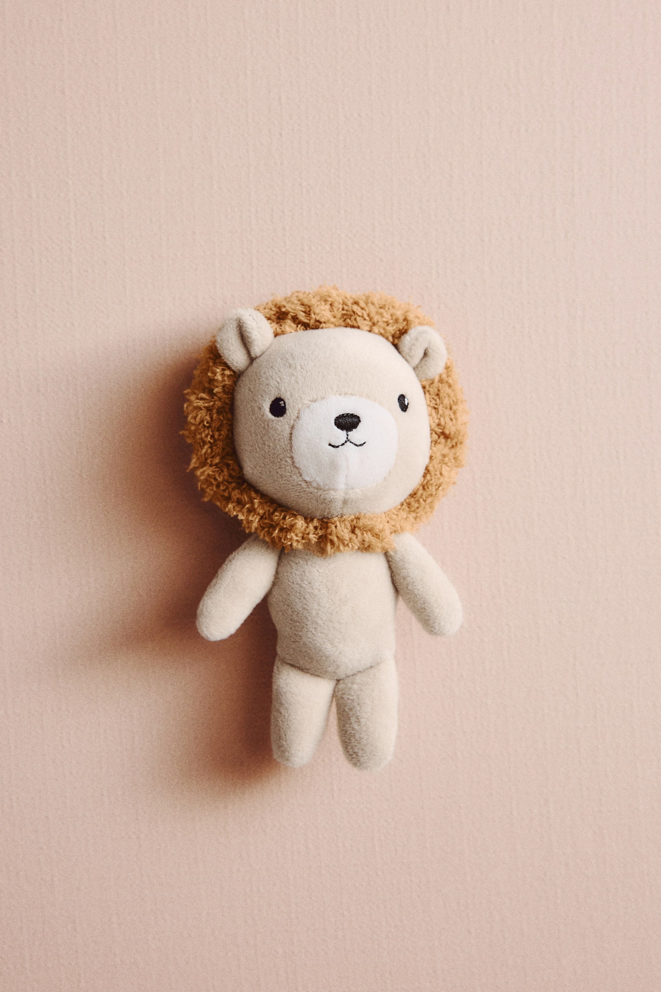 View larger image: A padded baby rattle, shaped like a lion, is facing front. It features a light cream soft pile body with a brown shaggy mane, and embroidered eyes, nose, and mouth.