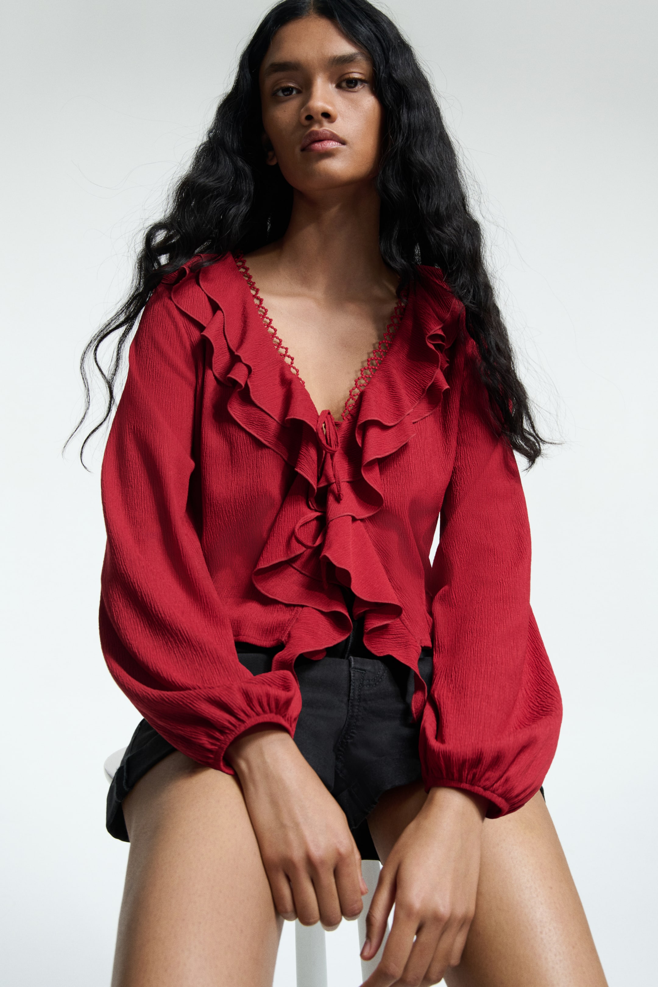 View larger image: A red textured blouse is featuring a V-neckline edged with a decorative picot trim, and cascading ruffles down the front secured by ties. It is presenting long, voluminous sleeves finished with gathered cuffs.
