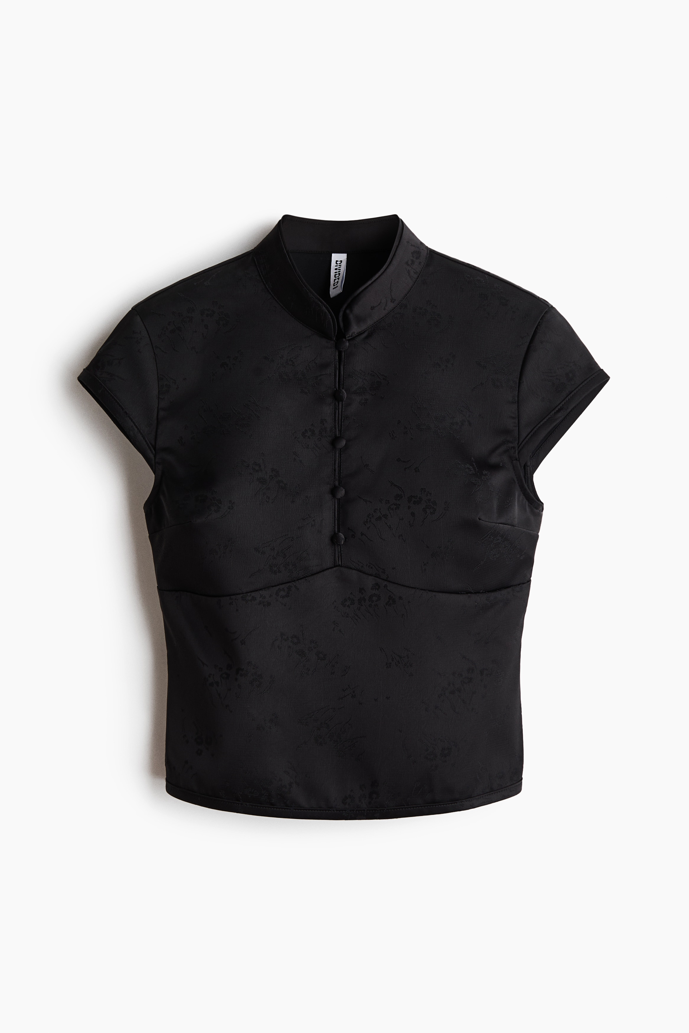 A black jacquard-weave top with a subtle sheen is being worn by the model, featuring a band collar with a buttoned placket, cap sleeves, and a curved seam below the bust. The top is paired with a matching black skirt in the same fabric.