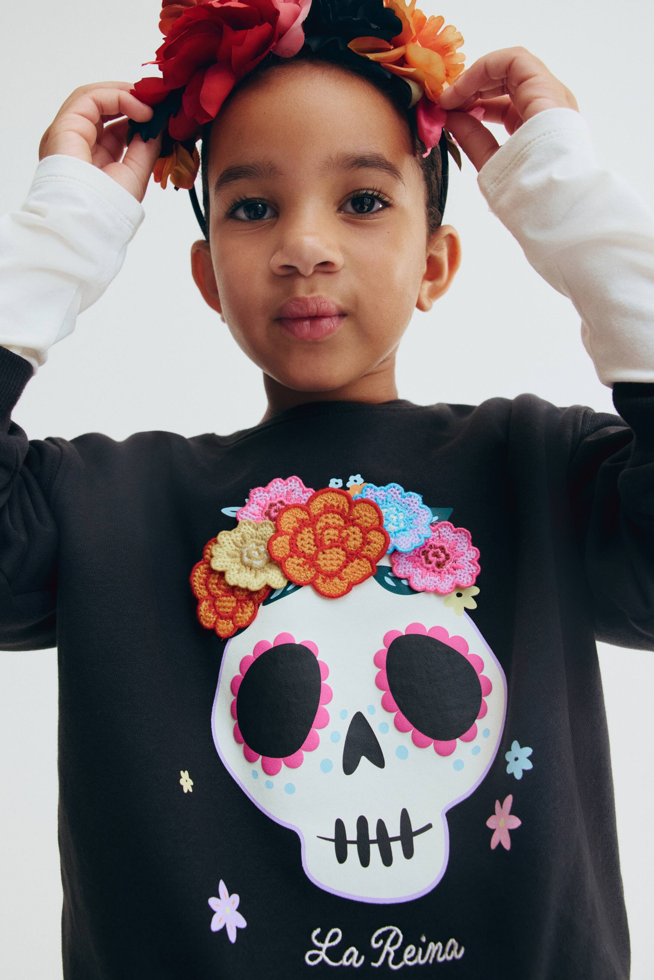 View larger image: A black crewneck fleece sweatshirt features a front-facing sugar skull graphic adorned with textured, colorful crocheted flower appliques. The design includes small printed pastel flowers around the skull and the white cursive text "La Reina" at the bottom, with white long-sleeve cuffs peeking out from under the ribbed cuffs.