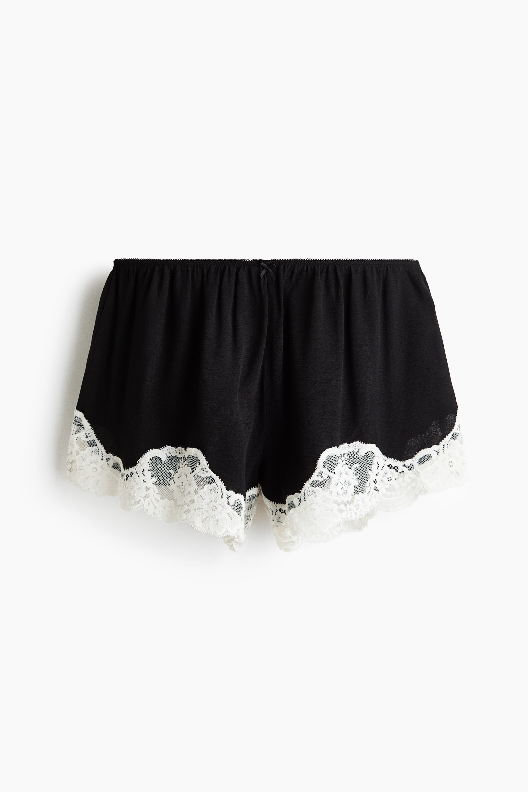 Ver imagem maior: Calções de pijama com acabamento em renda apresentam uma vista frontal, com um corpo principal preto e uma cintura elástica. Um pequeno laço preto adorna o centro da cintura, e uma larga renda branca com bordas recortadas decora as aberturas das pernas.