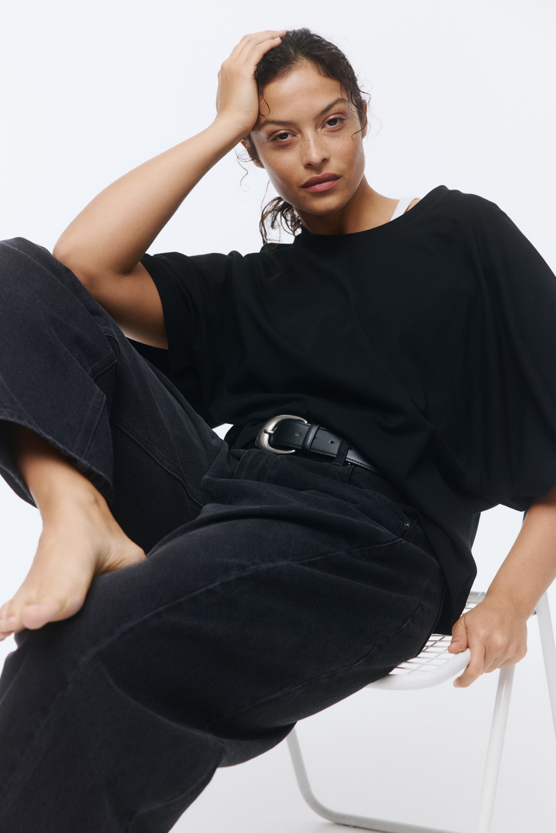 拡大画像を表示: 黒のオーバーサイズTシャツを着用しており、ラウンドネックと広がる袖が特徴です。ネックラインの下には、より明るい色の衣服が見えます。黒のリラックスフィットジーンズを履いており、ハイウエストで黒のベルトとシルバーバックルで締められています。