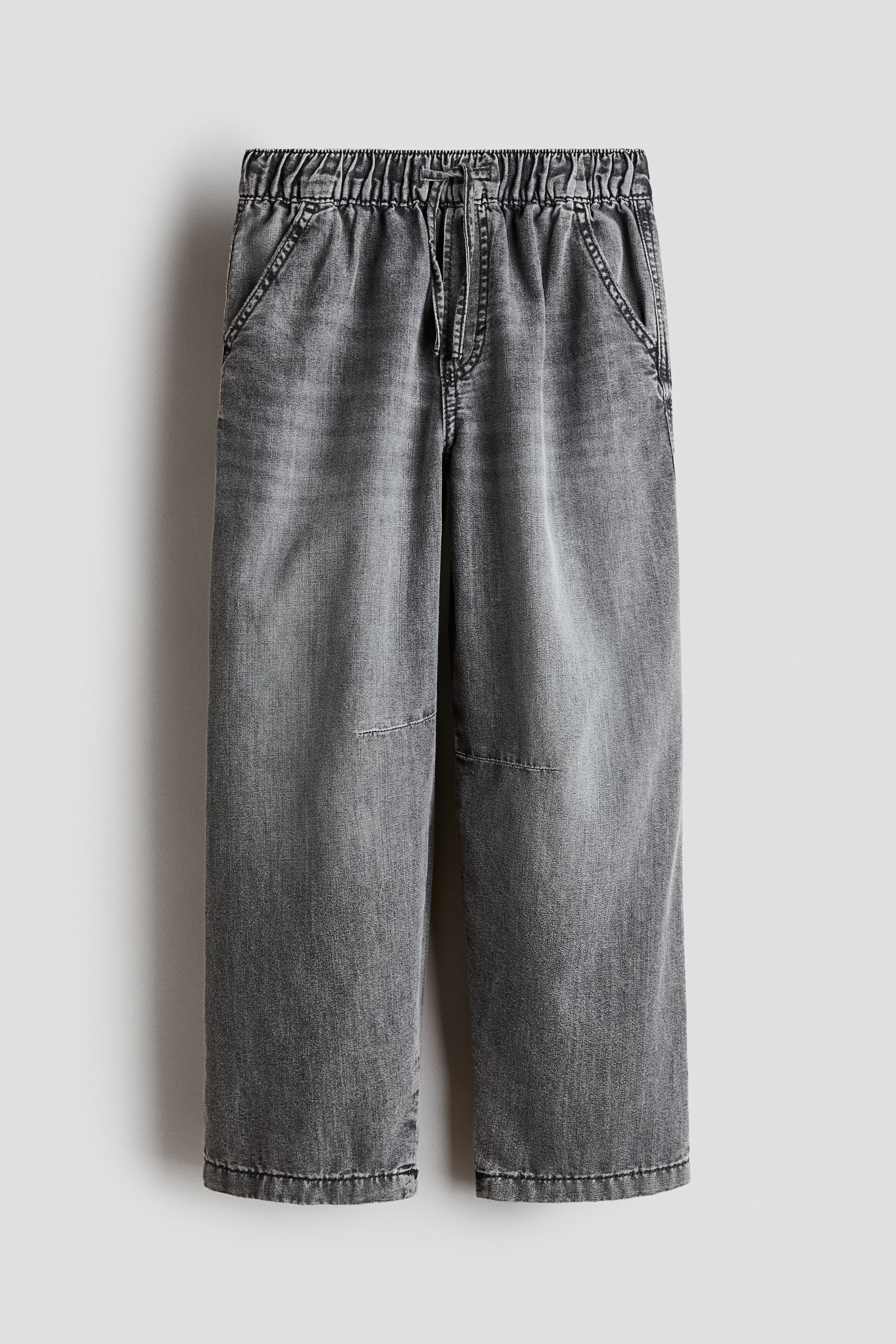 View larger image: A pair of grey washed denim pants is hanging front-facing, featuring a gathered elastic waistband with an integrated drawstring. The relaxed-leg pants display side slash pockets, horizontal knee seams, and a faded wash finish throughout.