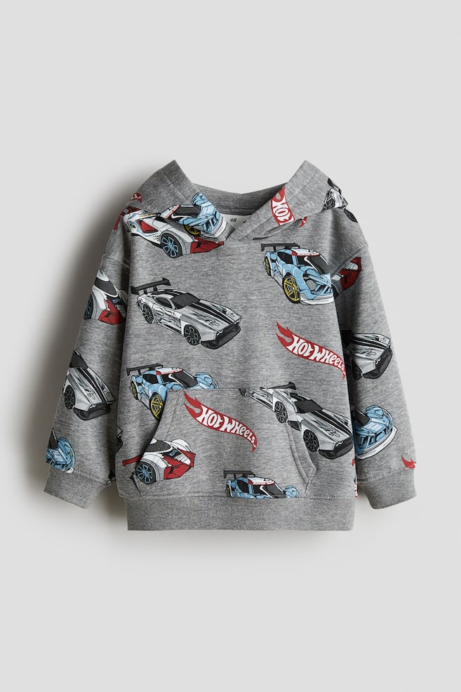 En grå hættetrøje vises FORAN, med et all-over print af farverige Hot Wheels-biler og logoer. Den er designet med en afslappet pasform, nedfaldne skuldre, lange ærmer, ribbede manchetter og kant, en foret omslagshætte og en stor kængurulomme. - 1