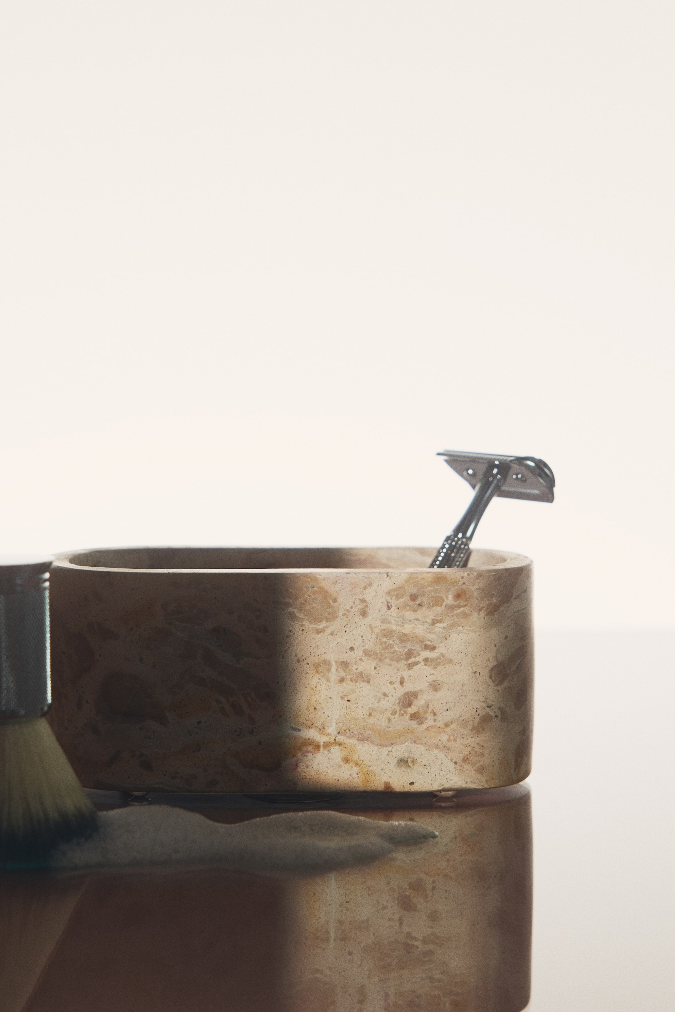 View larger image: An oval marble box in a beige, textured natural stone sits on a reflective surface, shown from the front without its lid. A silver safety razor rests inside the open container, and a shaving brush with lather is partially visible in the foreground.